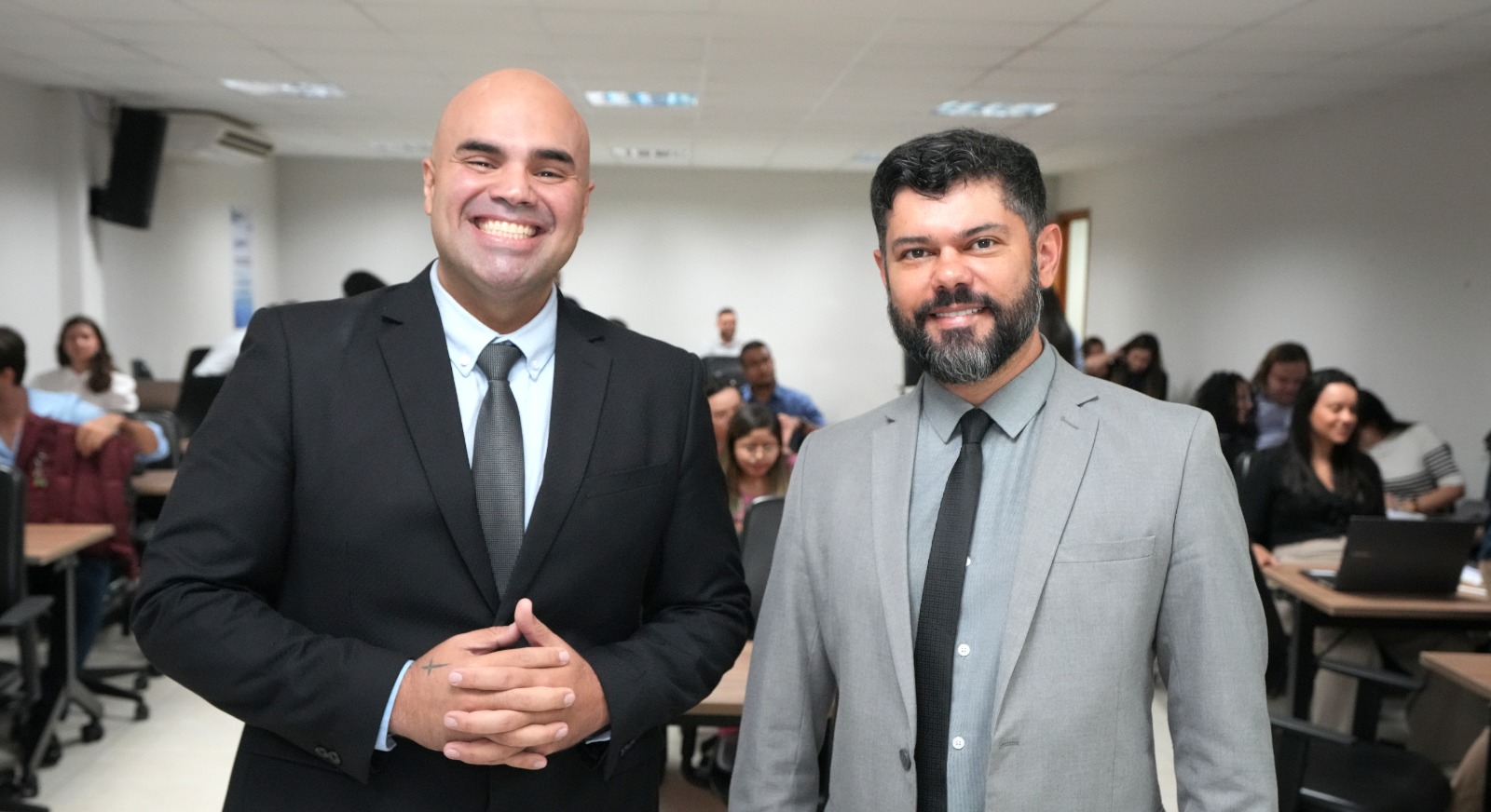 A imagem mostra dois homens no centro da cena, ocupando a maior parte do espaço visual. Eles estão vestidos com roupas formais. Ao fundo, há um grupo de pessoas em segundo plano, ligeiramente desfocadas, que estão participando de uma capacitação sobre sentença penal.