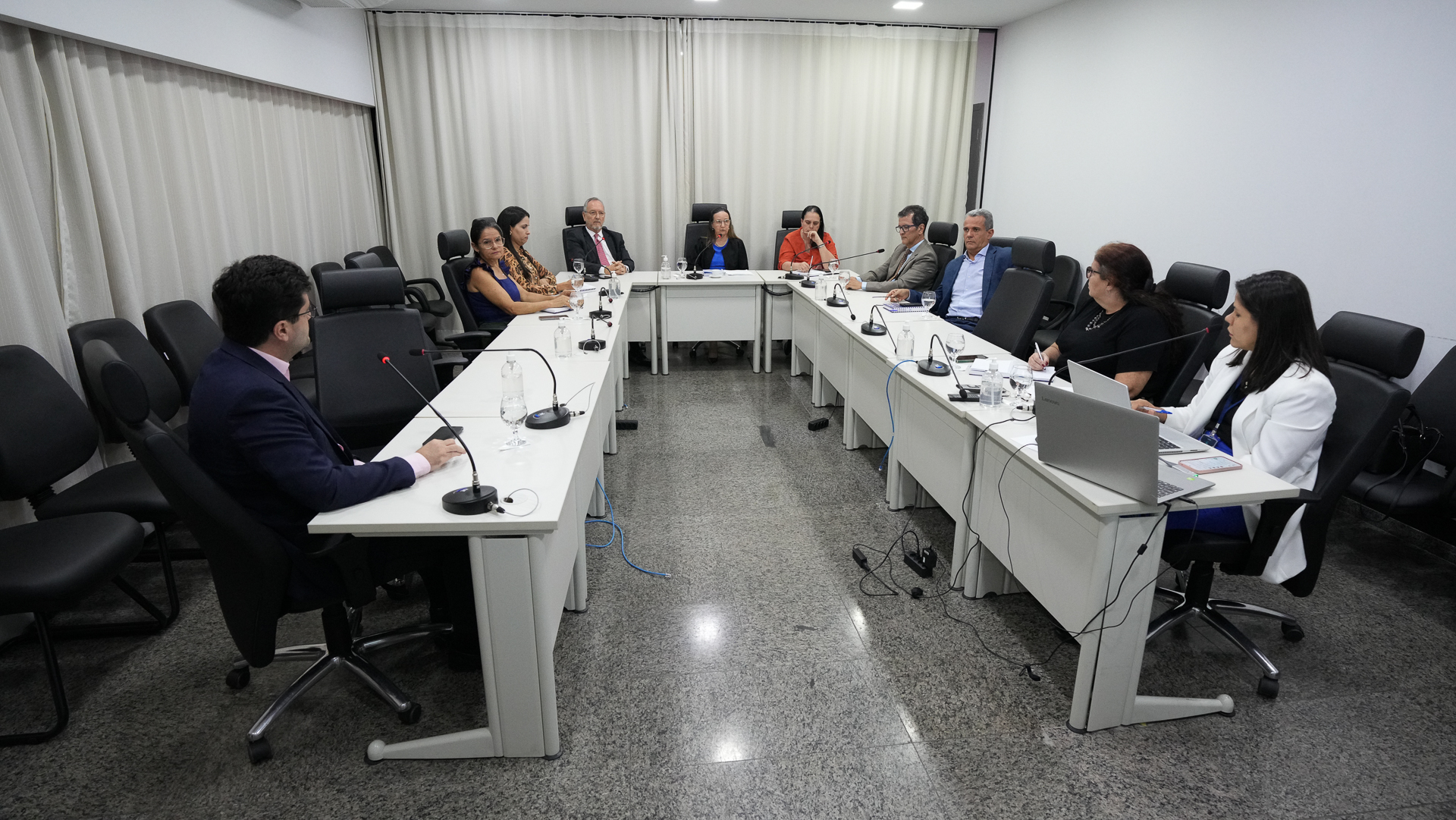 A imagem mostra uma reunião formal do Tribunal de Justiça do Tocantins (TJTO). O ambiente é bem iluminado, com luzes de teto brancas, cortinas claras e paredes neutras. Dez pessoas estão sentadas ao redor de uma mesa em formato de “U”, com microfones, blocos de anotações e copos de água à disposição, o que indica um encontro institucional ou estratégico.  Entre os participantes, há magistradas e magistrados, além de servidores, todos atentos à discussão. 