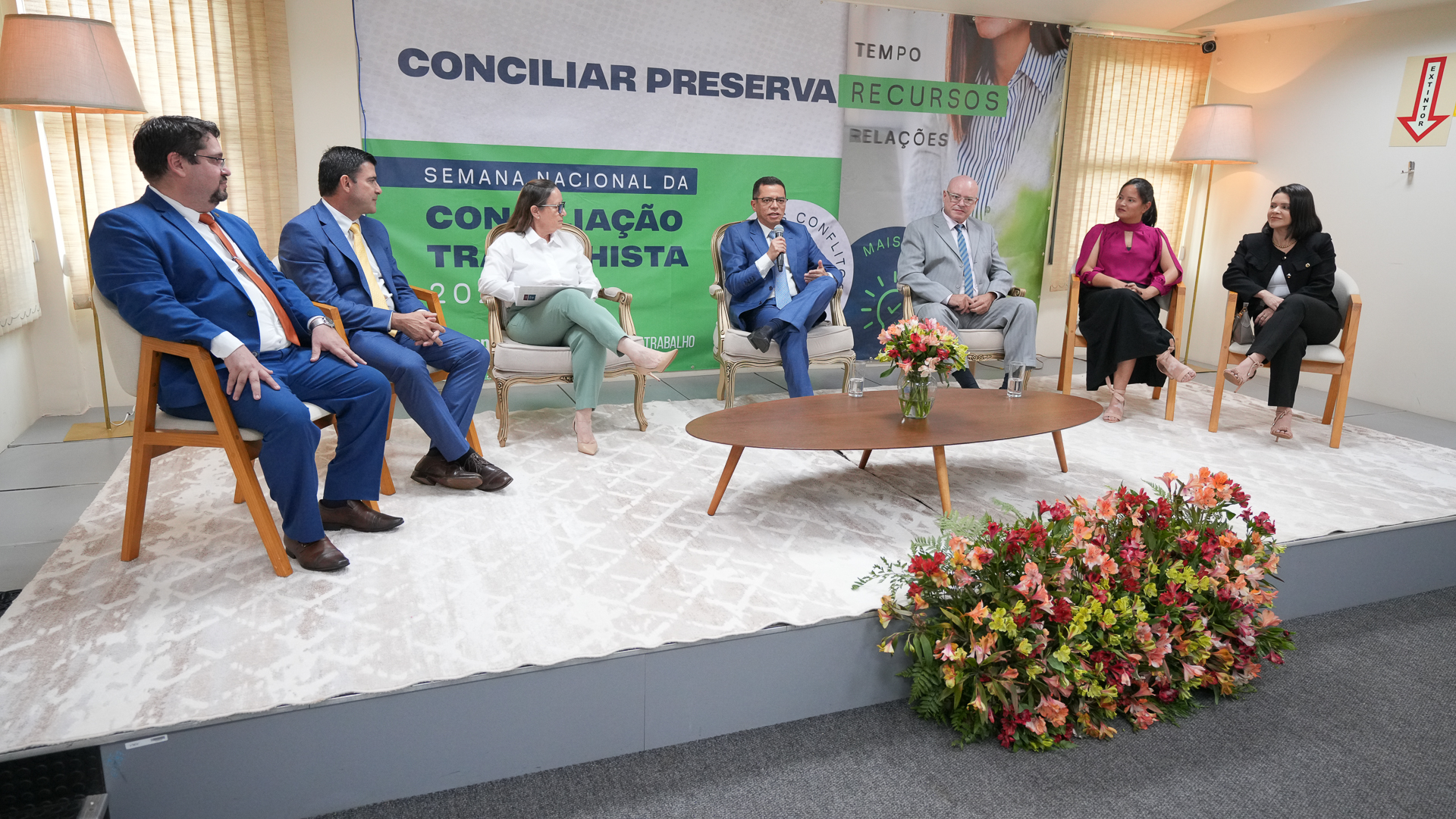 Na imagem aparecem sete pessoas sentadas em um formado de meia lua. No centro tem uma mesa de madeira com arranjo de flores. Os quatro homens e três mulheres estão participando do lançamento da semana nacional da conciliação trabalhista, em Palmas.