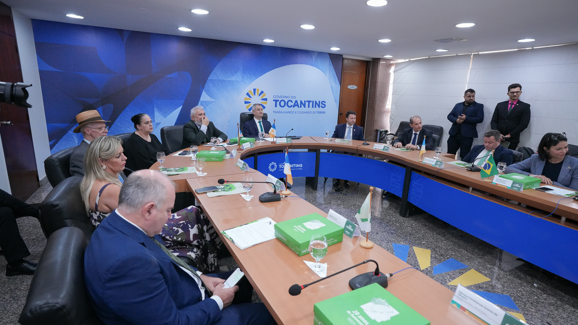 Na imagem aparecem 12 pessoas, dez sentadas em volta de uma mesa oval e 2 em pé do direito de quem olha. Todos estão participando de uma reunião do conselho nacional das defensoras e defensores-gerais. O encontro está sendo realizado na sala de reuniões do Palácio do Governo Estadual, em Palmas.