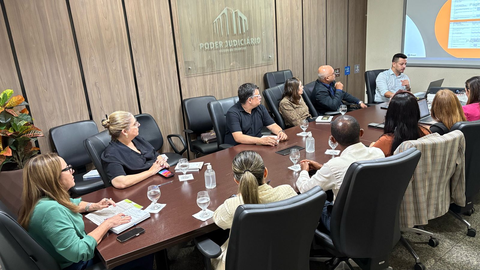 Homens e mulheres sentados em mesa de madeira, em sala de paredes amadeiradas e persiana bege