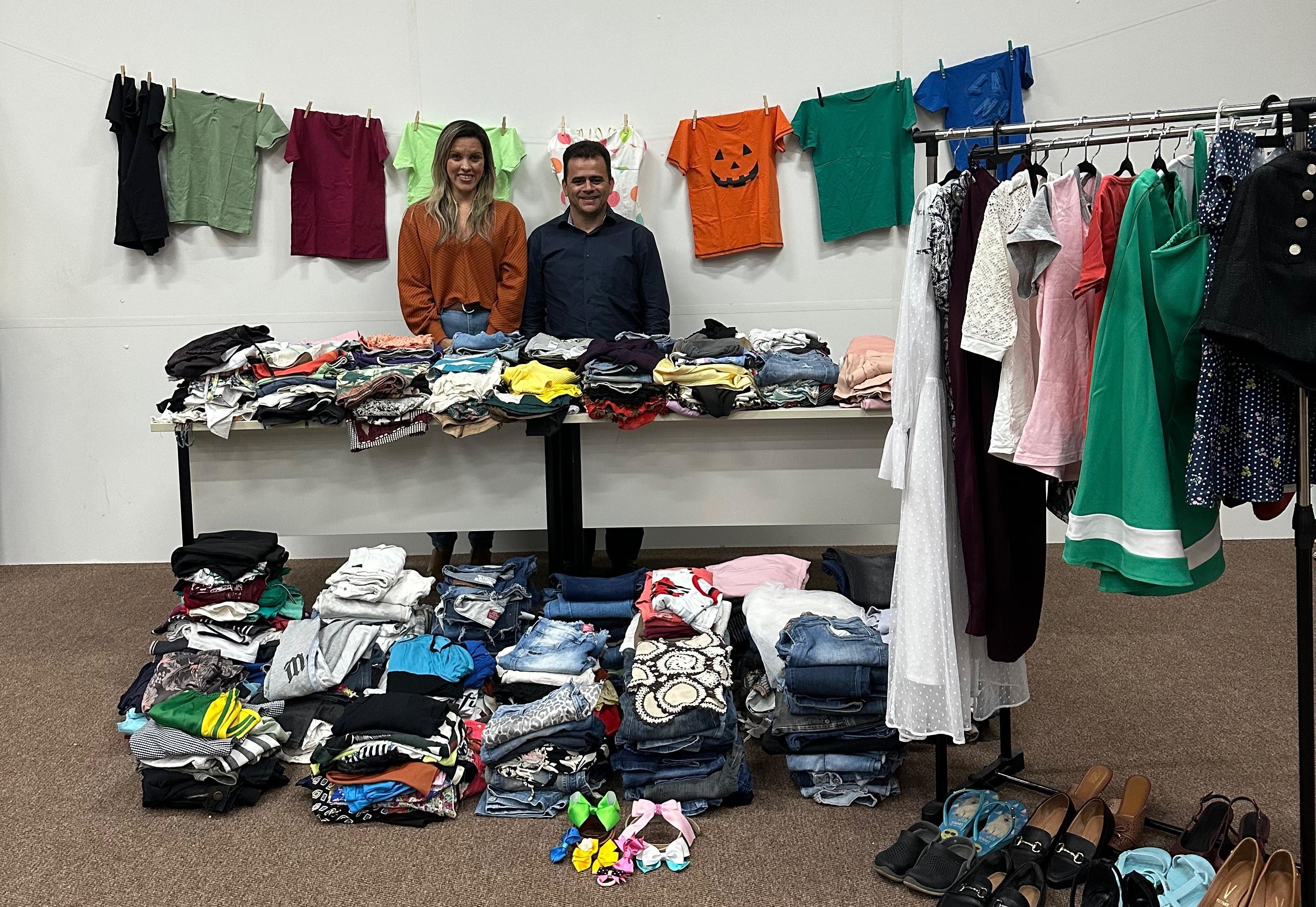 Uma mulher de calça jeans e camisa marrom está ao lado de homem de camisa azul marinho. Eles estão em meio a roupas e calçados, objeto de doações