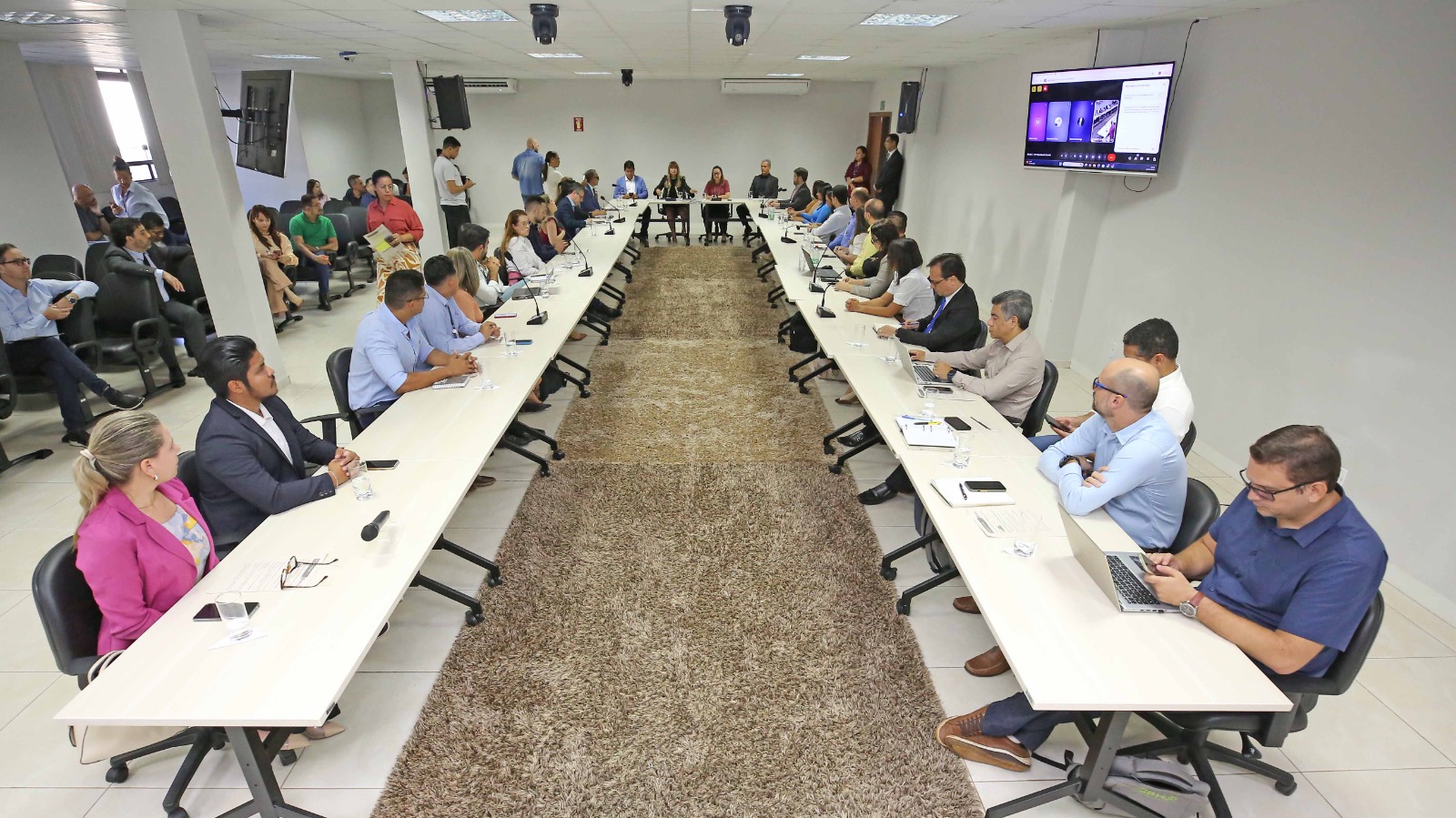 Na imagem aparece uma mesa no formato da letra 'U" dentro de uma sala bem espaçosa. Em volta da mesa estão sentadas várias pessoas. Elas estão participando de uma reunião do comitê estadual da saúde, onde foram apresentados os números da semana nacional da saúde.