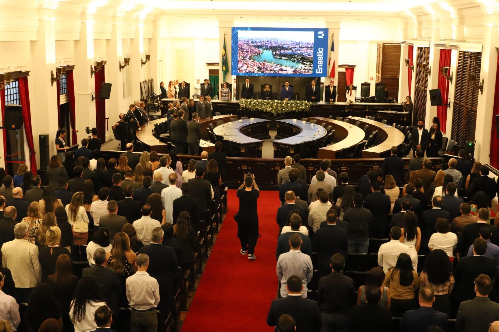 A imagem mostra muitas pessoas sentadas em fileiras em um salão amplo. O espaço central é ocupado por mesas em forma de semicírculo e algumas pessoas em pé.  