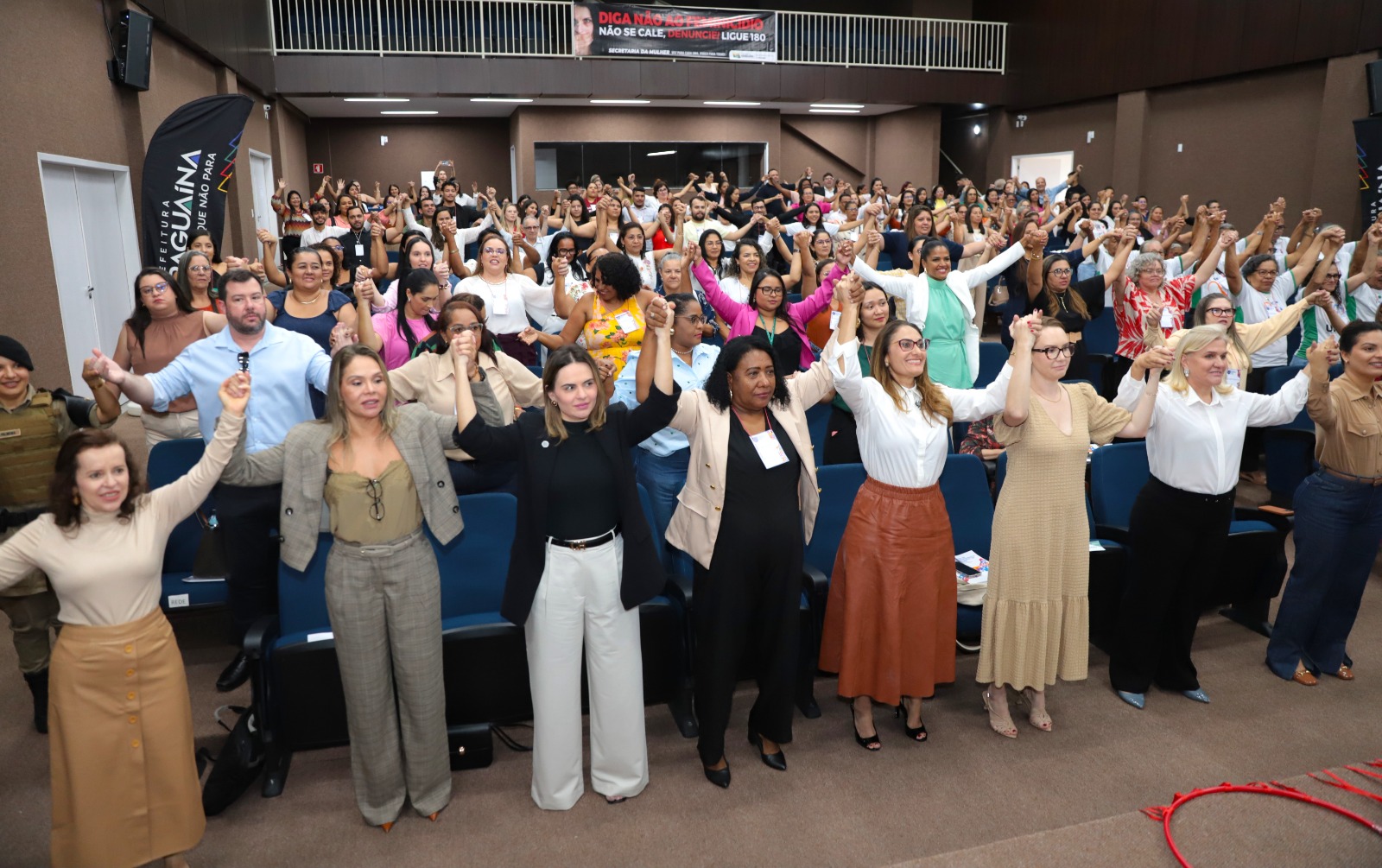 Imagem de um auditório cheio durante o "Fórum de Enfrentamento à Violência de Gênero", em Araguaína-TO. Participantes de diferentes idades, raças e gêneros estão de pé, de mãos dadas e com os braços erguidos, simbolizando união e força coletiva no combate à violência contra a mulher. Na primeira fileira, estão mulheres que aparentam ser autoridades ou organizadoras do evento. Ao fundo, uma faixa com os dizeres: "Diga não à violência. Não se cale. Denuncie. Ligue 180", da Secretaria da Mulher. O ambiente transmite engajamento, solidariedade e empoderamento.