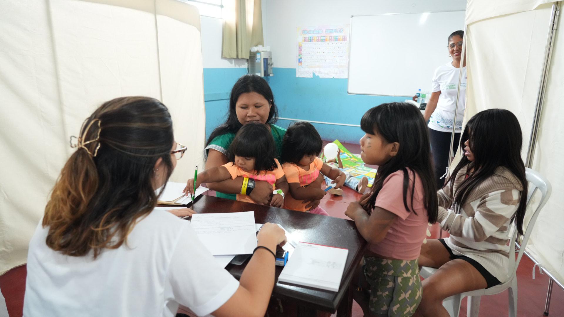 Mulher indígena sentada de frente com duas crianças gêmeas no colo e outras duas maiores do lado sendo consultada por uma médica que aparece de costa. 