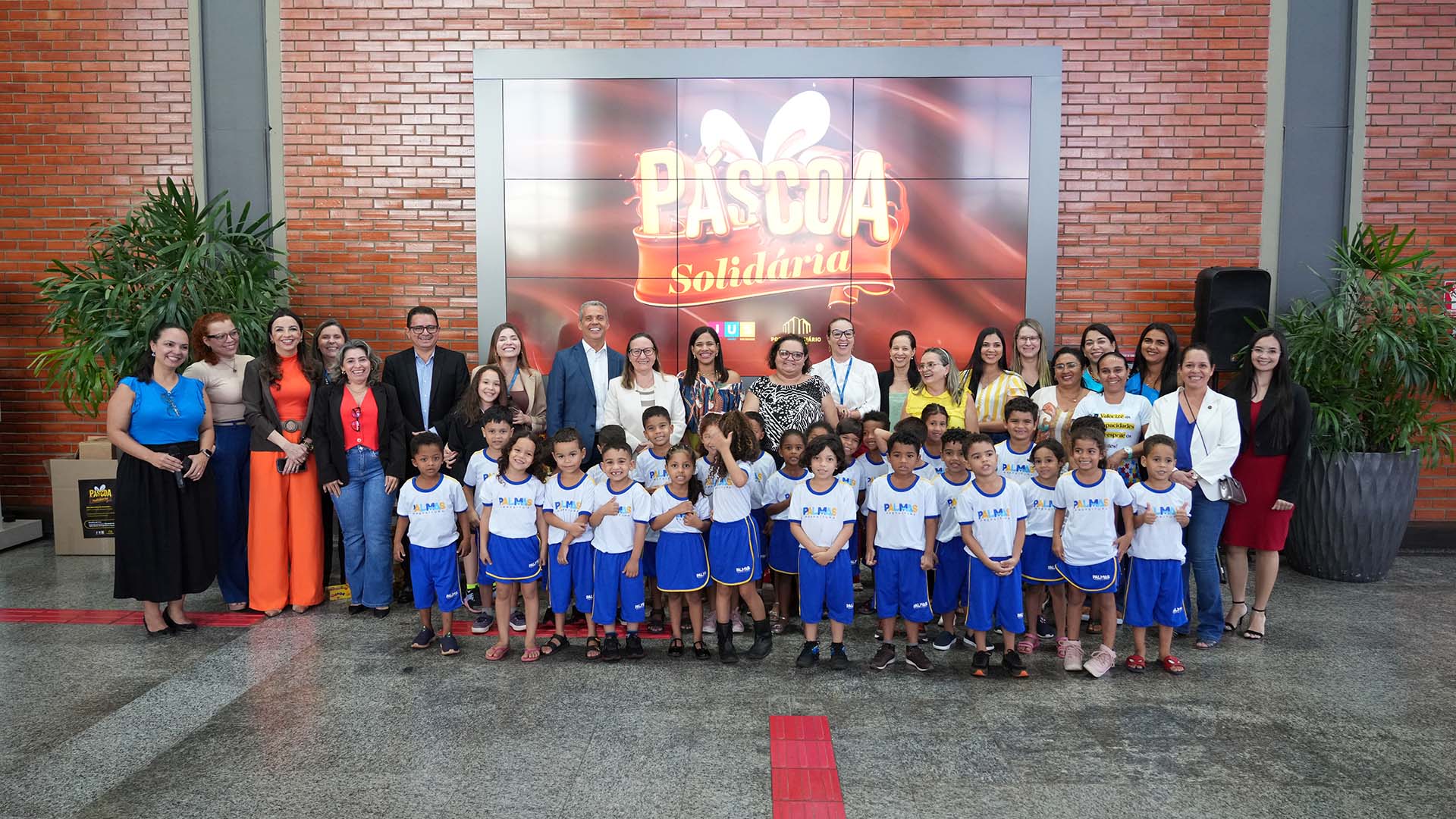 Foto de um grupo de adultos e crianças durante evento de encerramento da campanha Páscoa Solidária. As crianças no centro da imagem e usam uniformes de escola azul, com detalhes em branco. Há um grande painel de fundo com um anúncio da Páscoa Solidária.