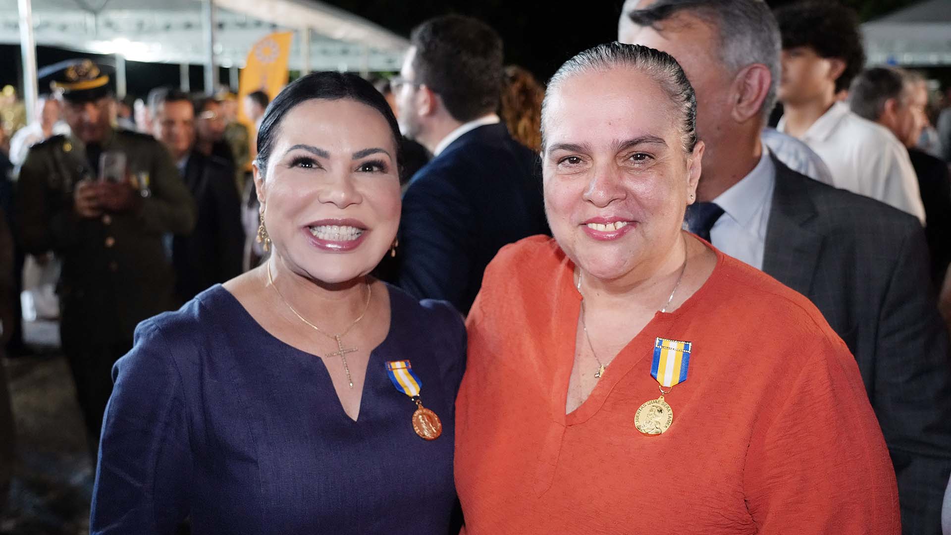 Imagem mostra a vice-presidente do TJTO e a juíza Edilene em pé, sorrindo para a foto, durante o evento; a desembargadora Jacqueline está à direita, de roupa laranja, e a juíza à esquerda de roupa azul; ambas usam medalha recebida no peito, lado direito. 