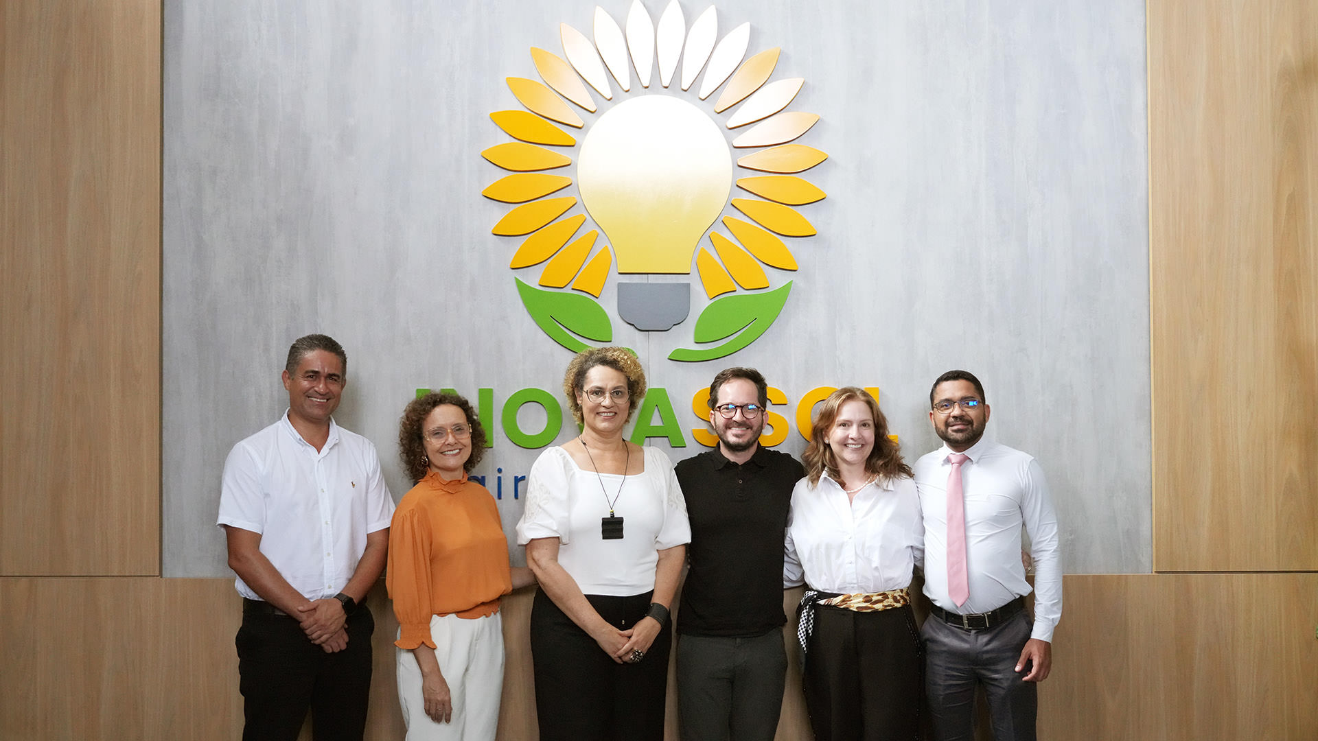 Imagem mostra um grupo de pessoas, sendo três homens e três mulheres, todos posicionados em uma fileira, um ao lado do outro, na frente de um fundo de parede com um logotipo colorido de uma lâmpada no centro de um girassol.