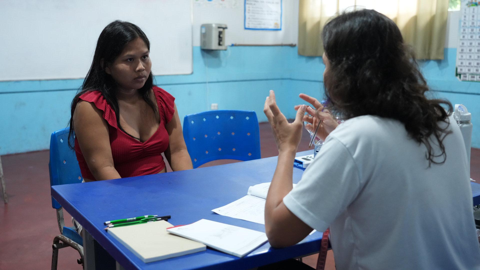 Mulher indígena grávida sentada de frente para outra mulher, médica, numa sala de consultas