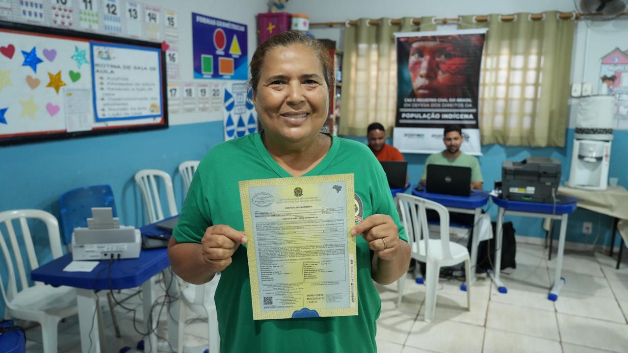 Imagem mostra uma mulher sorridente, de roupa verde, segurando um documento com ambas as mãos à frente de si. Ao fundo aparecem outras pessoas em atividades relacionadas a computadores e trabalho em mesas na sala. 