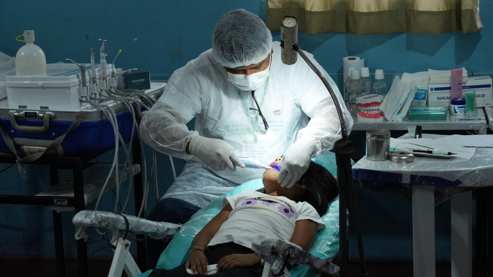 Imagem mostra um dentista com jaleco branco, touca e máscara, atendendo uma criança indígena que está sentada na cadeira de dentista