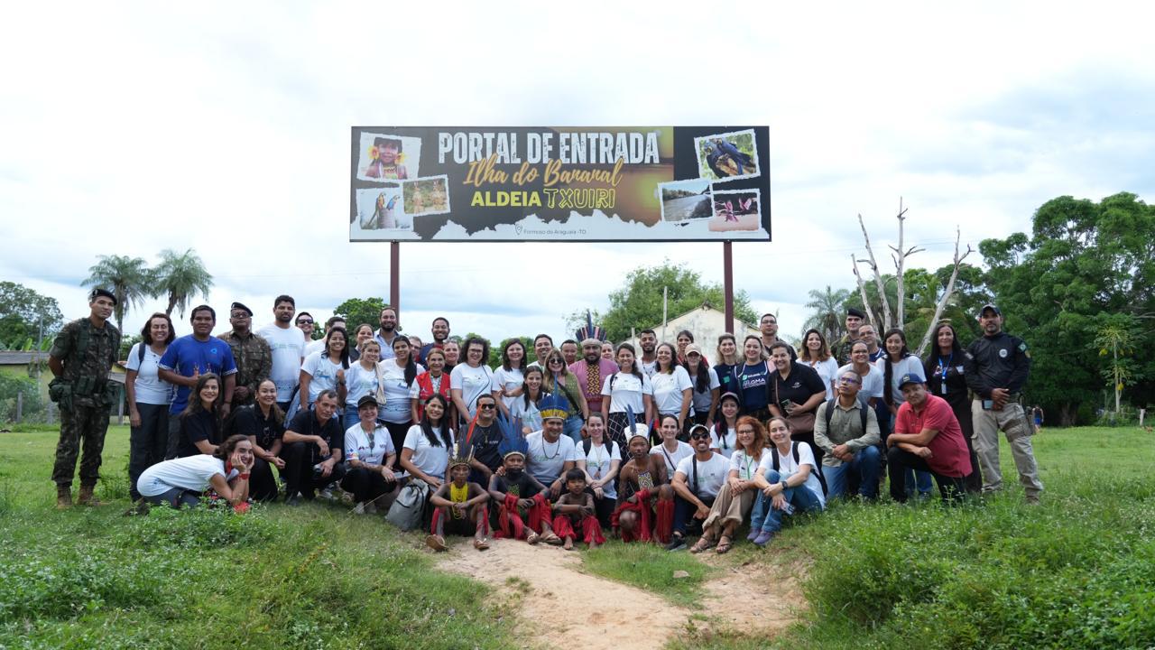 Imagem mostra um grupo grande de profissionais que trabalharam na Semana Nacional de Saúde posicionados em frente à placa na entrada da Aldeia Txuirí, na Ilha do Bananal. 