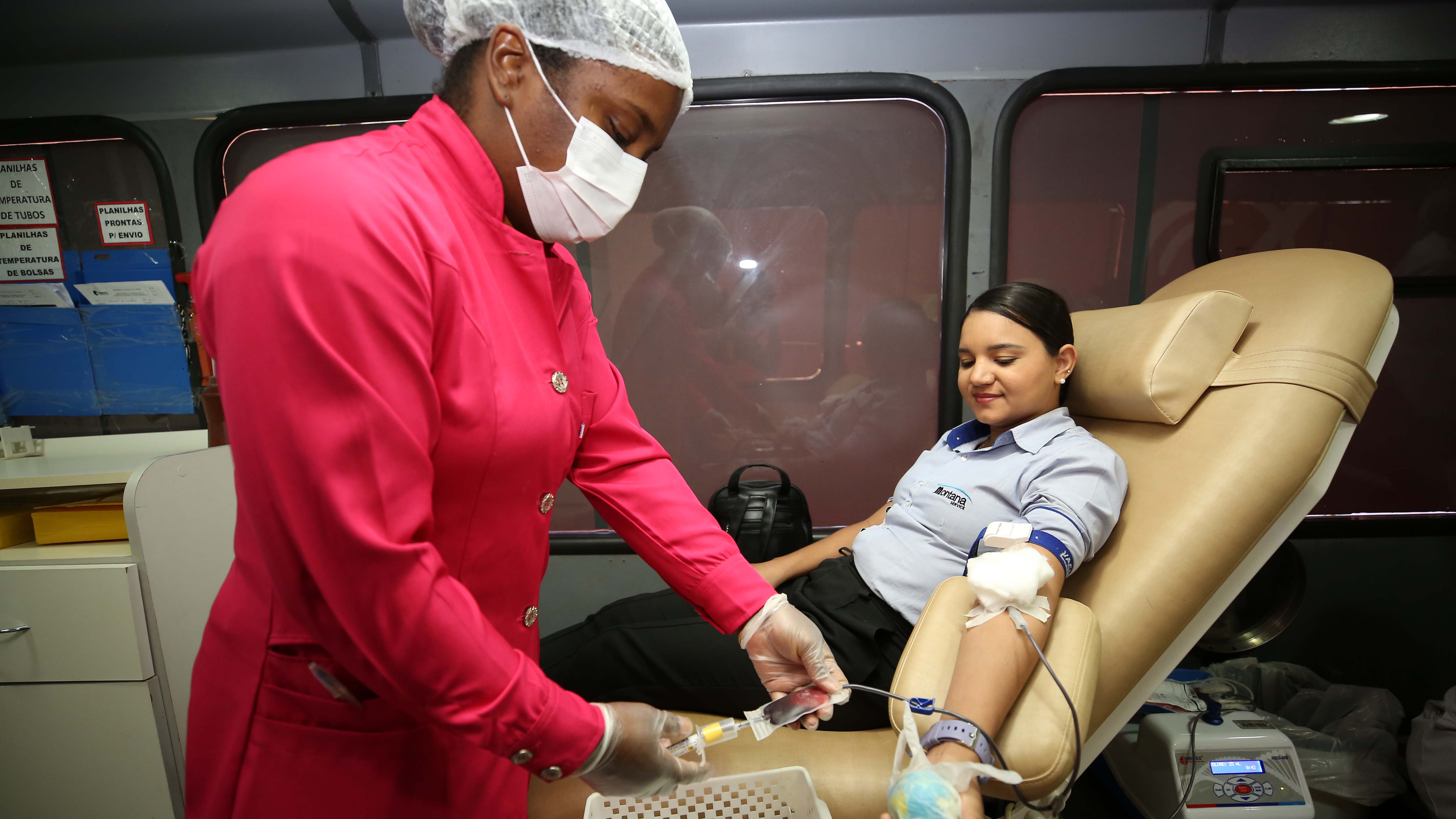Em um ônibus do Hemocentro, servidora sentada em uma poltrona doa sangue; ao seu lado uma profissional de saúde do Hemocentro, de touca, máscara e jaleco vermelho