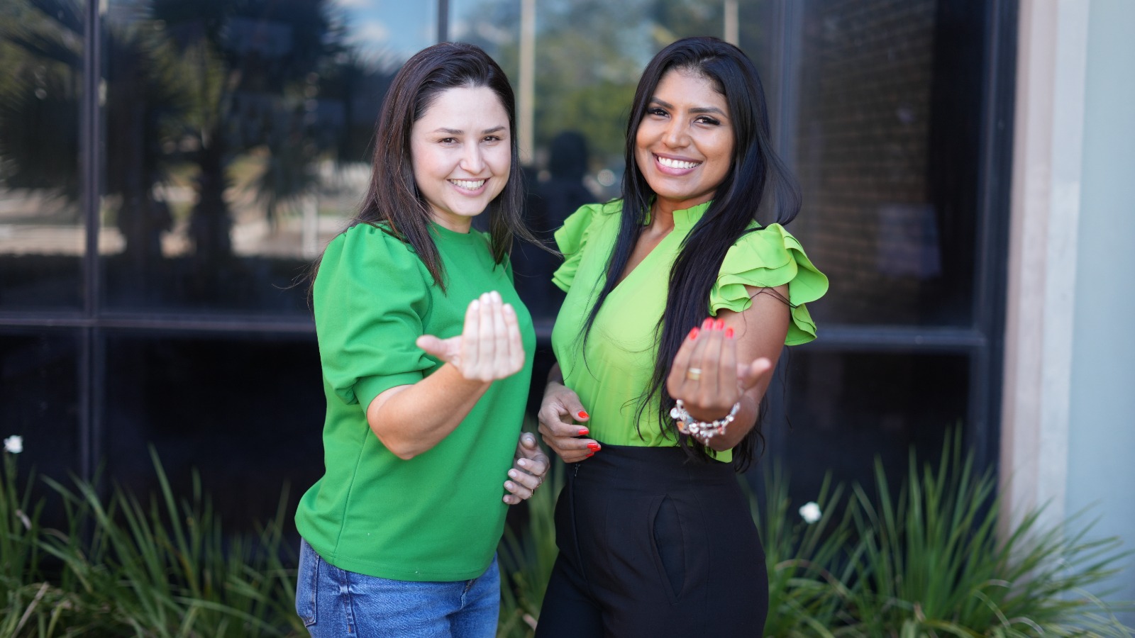 uas mulheres sorridentes, vestindo tons de verde, posam para a câmera em frente a um prédio com janelas espelhadas e plantas ao fundo. Ambas fazem com a mão o gesto simbólico do 'vem comigo', com os dedos unidos apontados para frente. A mulher à esquerda veste uma blusa verde e calça jeans, enquanto a da direita usa uma blusa verde-limão com babados nas mangas e calça preta. O gesto reforça a ideia de convite ou mobilização, possivelmente relacionada a uma campanha institucional.
