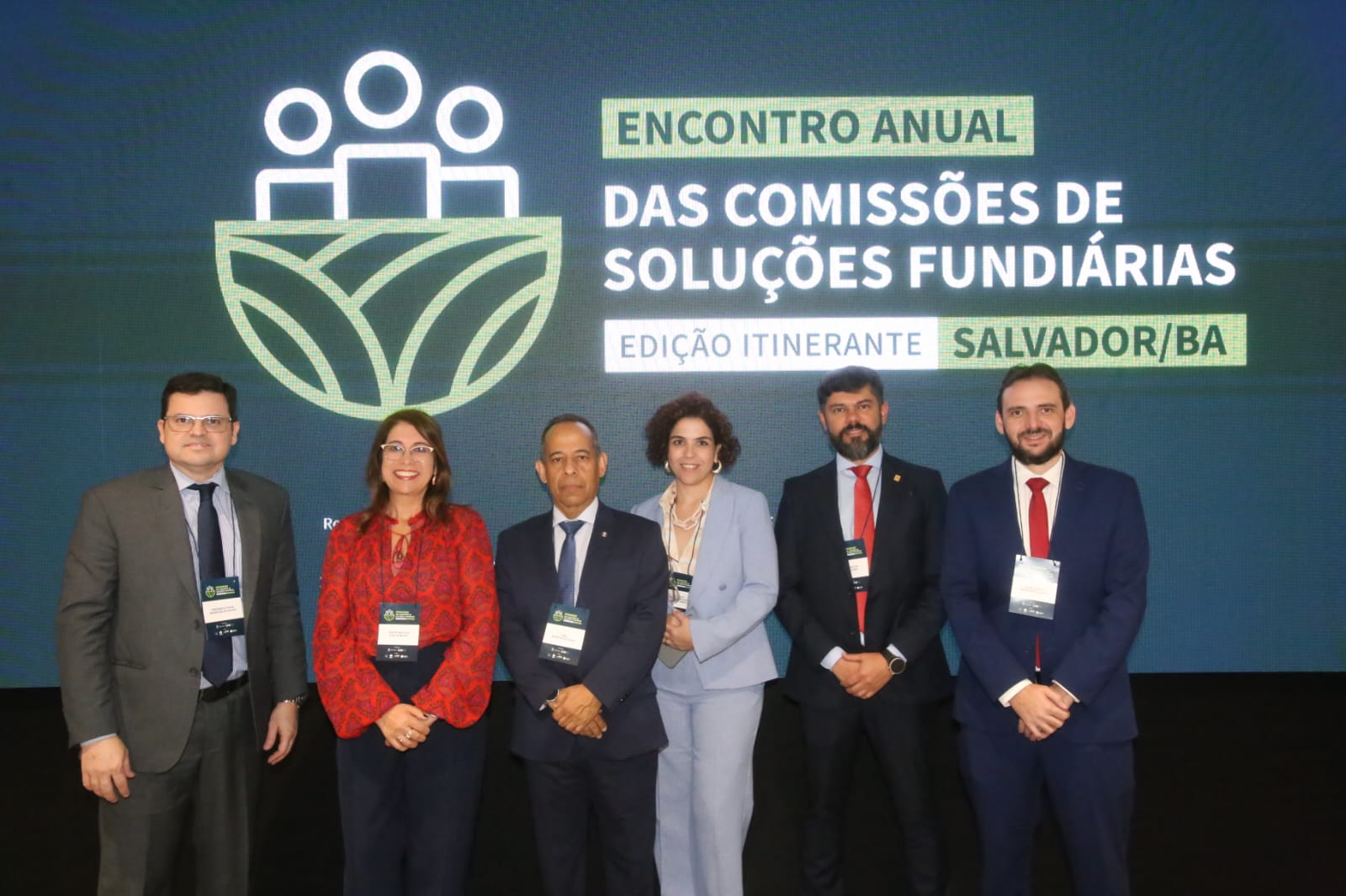 Imagem mostra seis pessoas com roupas formais em pé; são quatro homens e duas mulheres; ao fundo um banner do evento com  o nome Encontro Anual das Comissões de Soluções Fundiárias