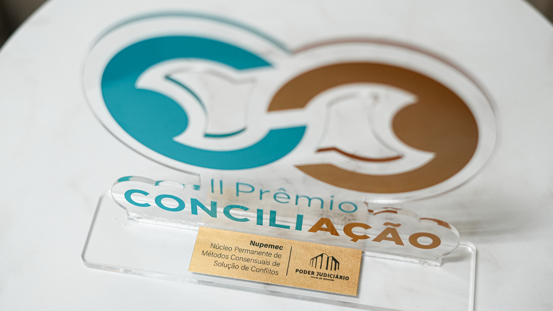 foto de um troféu do II Prêmio Conciliação em material acrilico transparente com desenho e fontes nas cores azul e marrom; na base uma plaquinha dourada com o nome Nupemec e Poder Judiciário do Tocantins