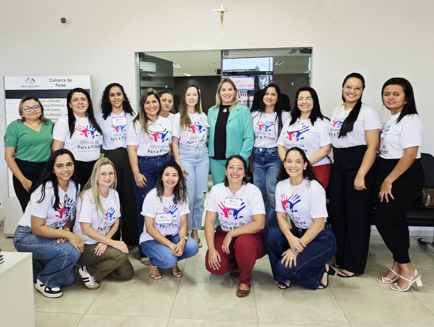 Dezesseis mulheres posam para  foto; 11 estão em pé e  cinco agachadas; a maioria usa camisetas brancas com logo e nome: Oficina de Pais e Filho  