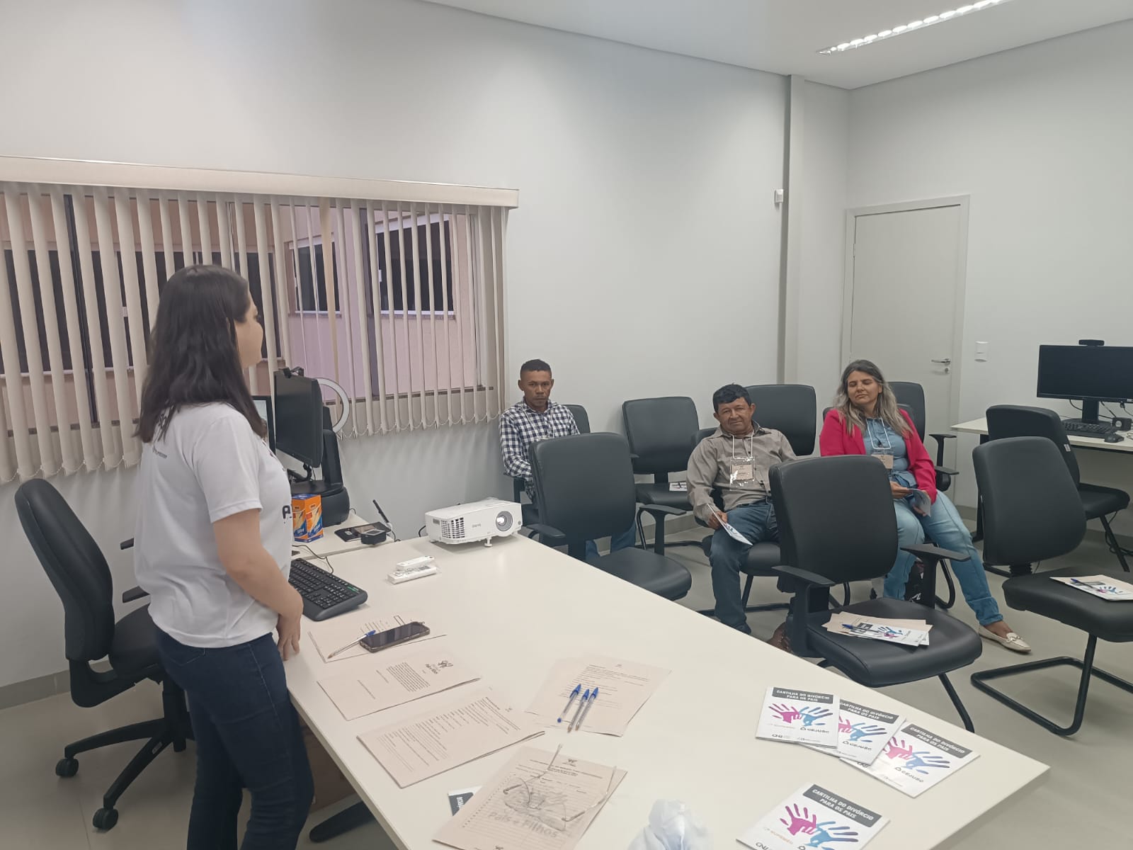 Imagem de uma sala com várias cadeiras e algumas pessoas sentadas olhando para uma mulher que está em pé de camiseta branca e calça escura falando de frente para elas, à frente de uma mesa branca