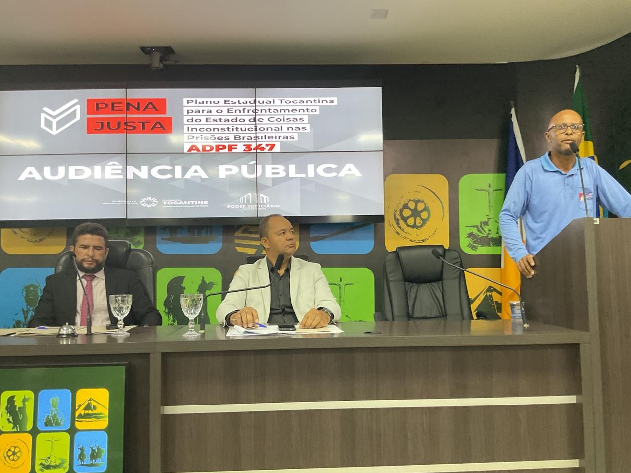 Imagem mostra duas autoridades sentadas, à esquerda, observando um representate da sociedade falando ao microfone, à direita