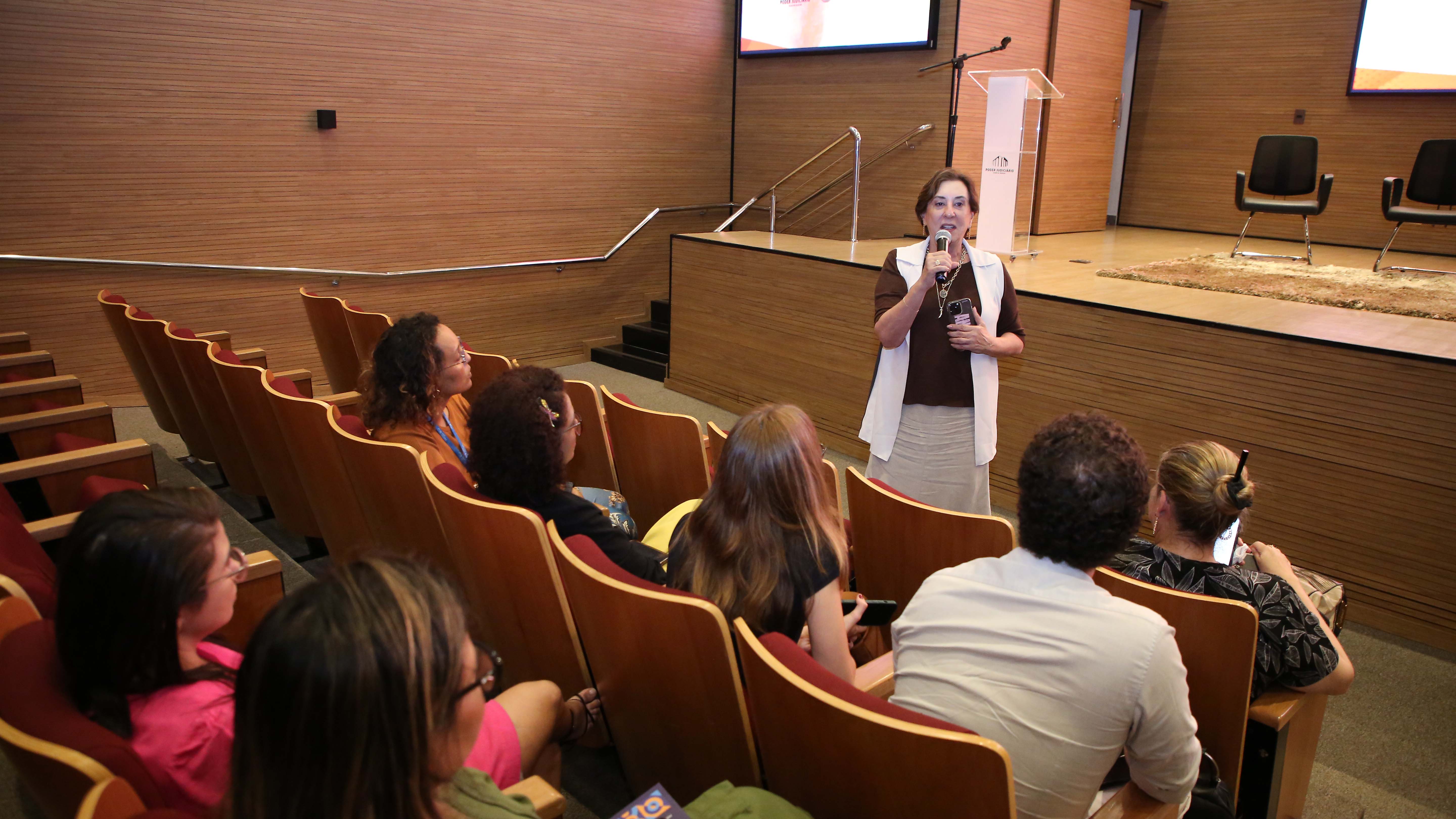 Mulher branca, alta, magra de cabelos curtos, vestindo saia bege e blusa preta com colete branca, fala ao microfone para plateia sentada em auditório todo em acabamento amadeirado