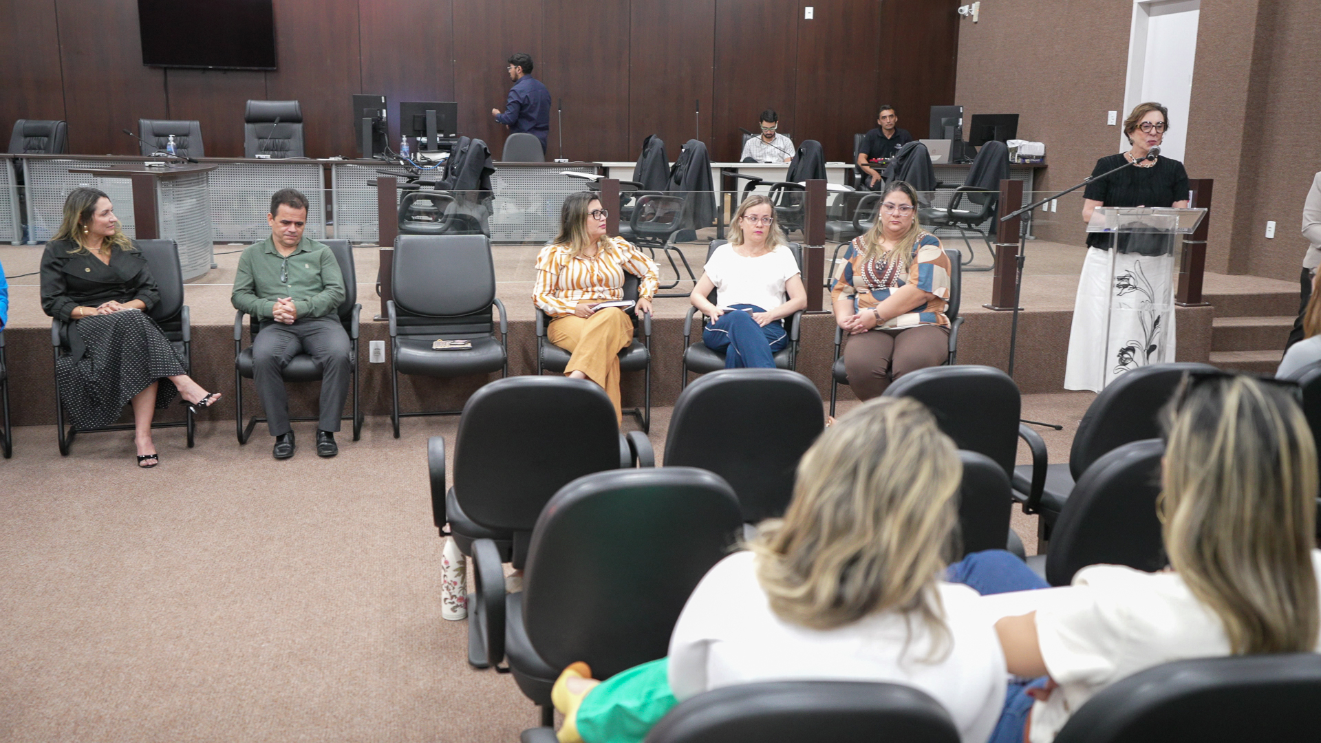 A imagem mostra uma sala de audiências com várias pessoas sentadas, enquanto uma mulher fala em um púlpito.