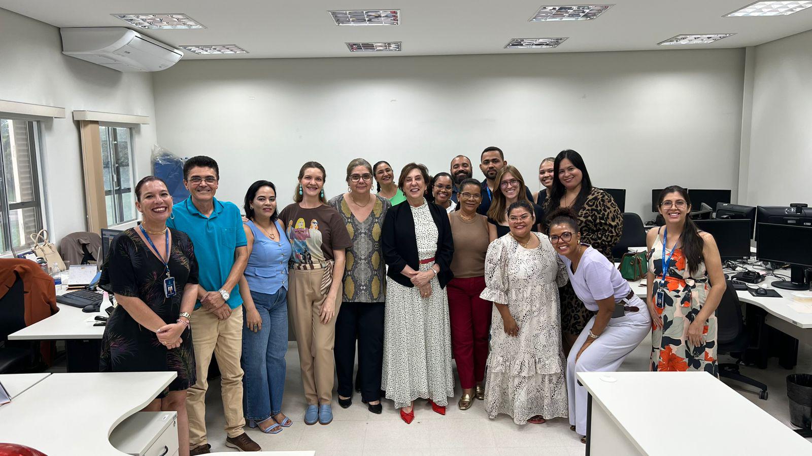 Foto em uma unidade do Fórum de Palmas com 15 pessoas alinhadas e sorrindo para a câmera. Elas formam uma fileira única no centro da sala, que tem mesas brancas, computadores e janelas ao fundo. O grupo é diverso em gênero, idade, altura e estilo, usando roupas sociais ou casuais; o clima é amistoso e descontraído.
