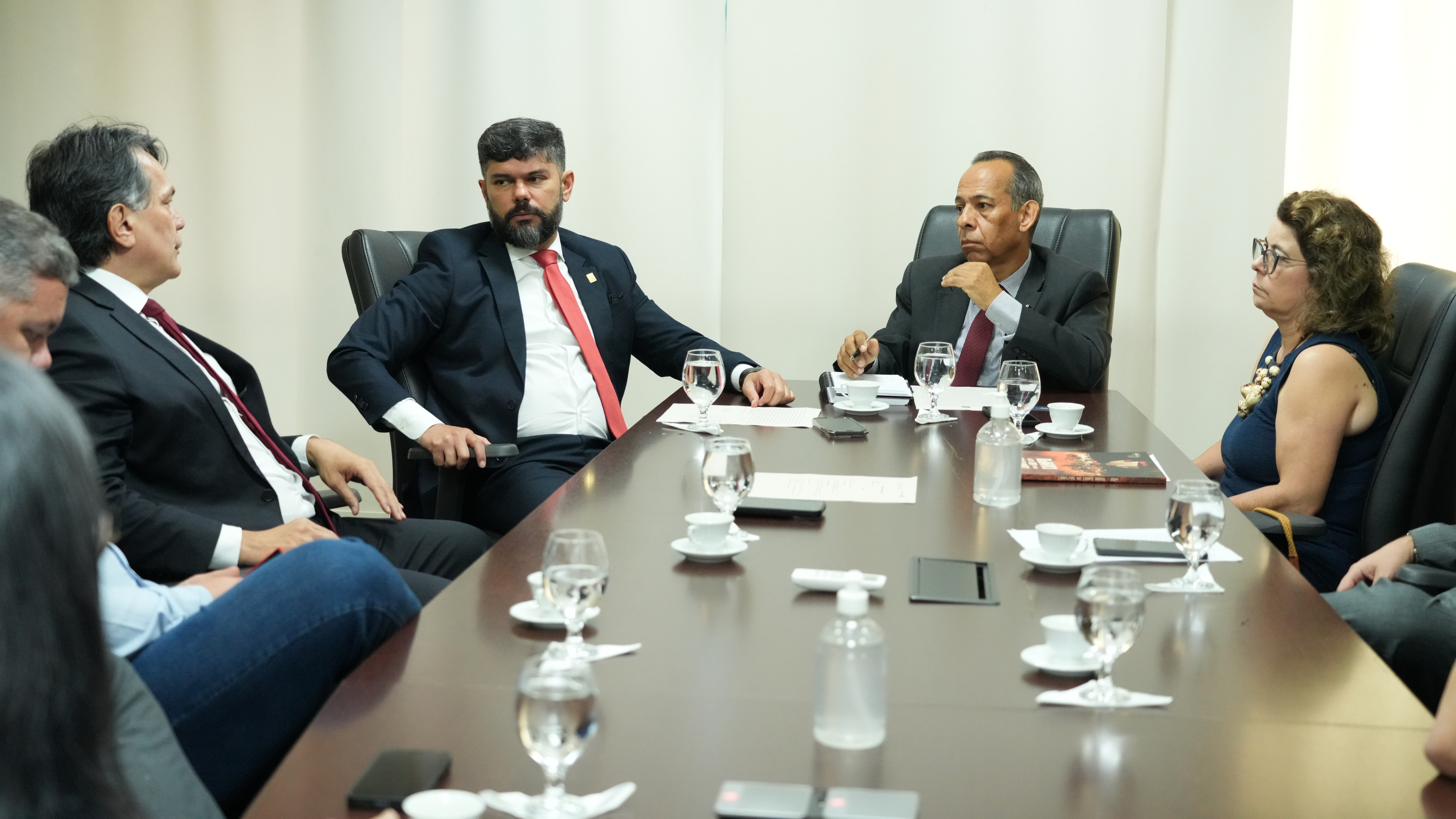 Homens de ternos escuros e uma mulher de camisa preta sentados à mesa de madeira em sala com cortina bege ao fundo