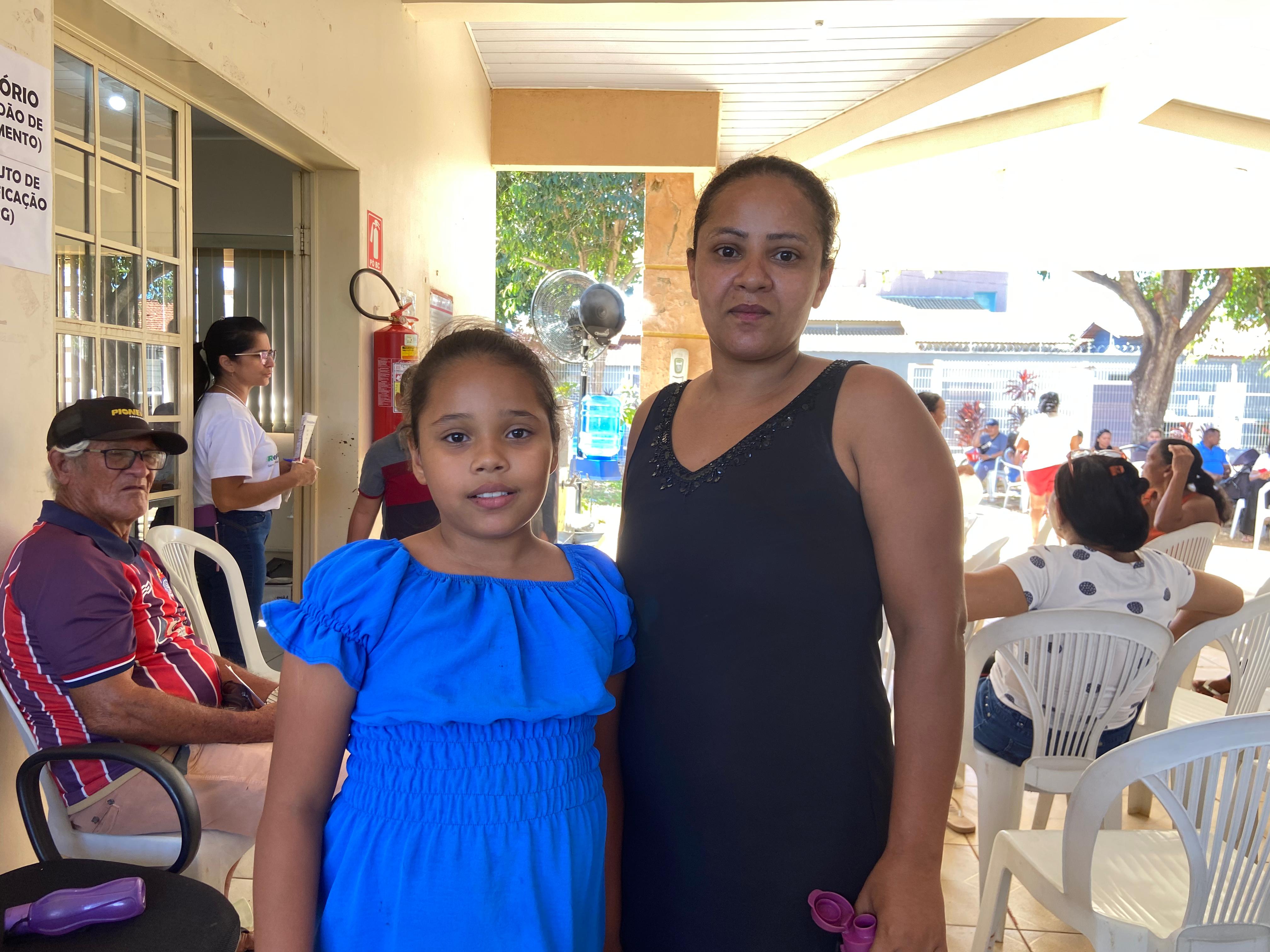 A imagem mostra um grupo de pessoas reunidas em um espaço aberto, sob uma estrutura coberta. Há um grupo de cadeiras de plástico branco dispostas no local. A mulher adulta e a menina estão no centro da imagem, em primeiro plano, e outros indivíduos estão visíveis em segundo plano. A mulher está vestindo um vestido preto sem mangas e a menina um vestido azul.