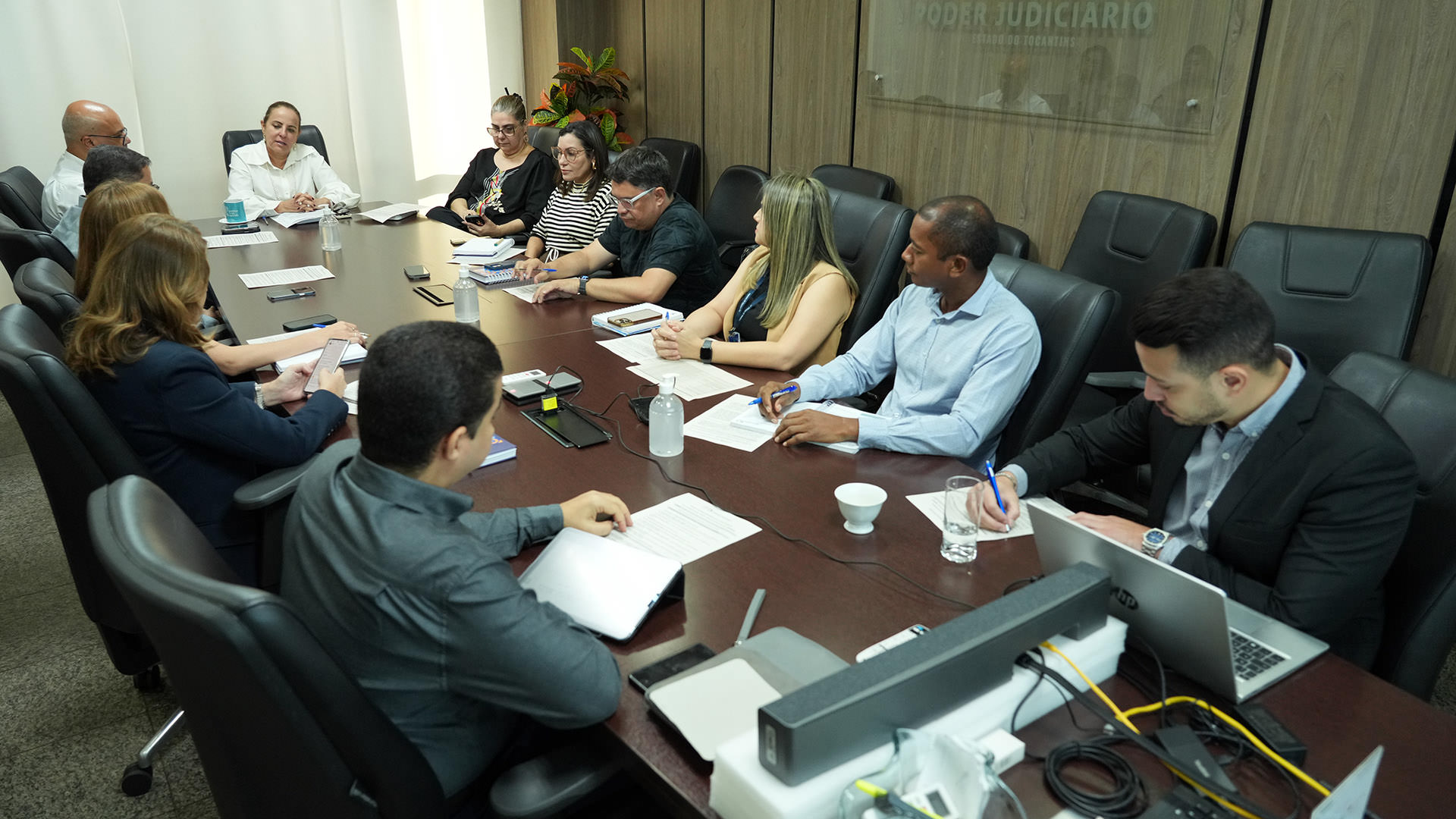 Homens e mulheres sentados ao redor de mesa de madeira durante reunião em sala de parede revestida de madeira. Ao fundo, parede com cortina bege