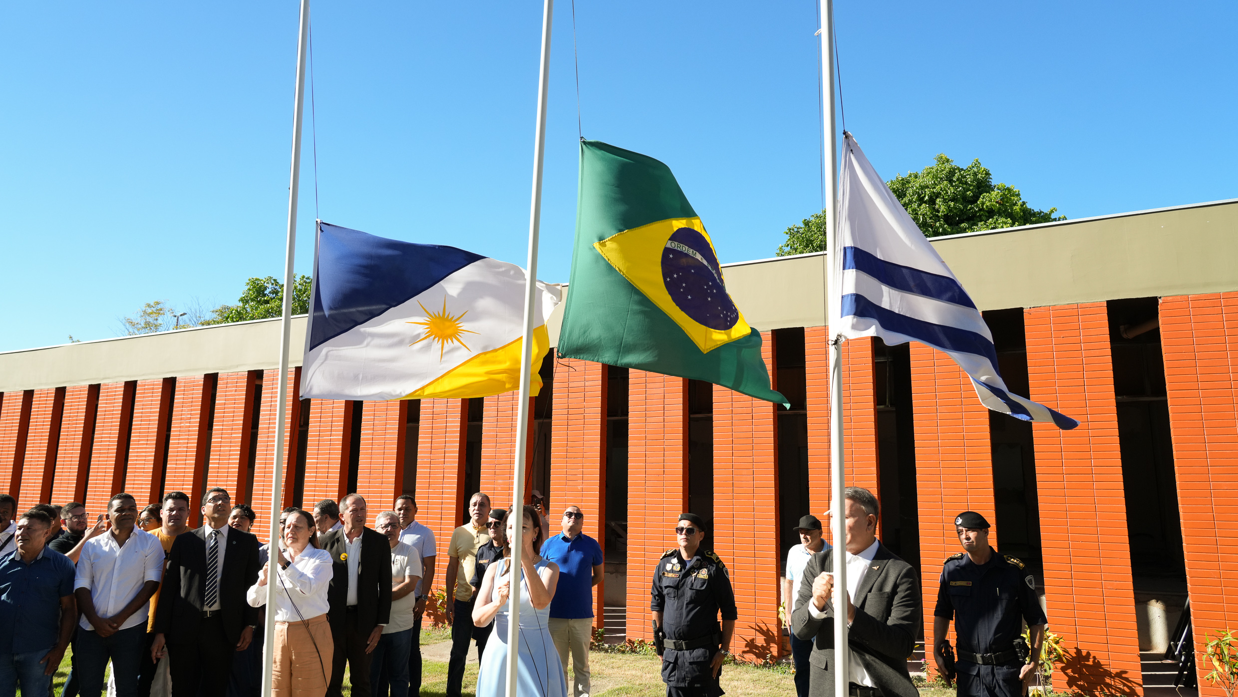 Cerimônia de hasteamento de bandeiras em frente a um prédio de tijolos aparentes, com paredes em tom laranja e detalhes verticais. Três bandeiras estão em destaque ao centro, hasteadas a meio mastro: a bandeira do Tocantins (à esquerda), a bandeira do Brasil (ao centro) e a bandeira de Palmas, capital do Tocantins (à direita). Um grupo de pessoas acompanha a cerimônia, incluindo homens de terno e policiais militares fardados. Duas pessoas estão posicionadas à frente, cada uma hasteando uma das bandeiras laterais. O céu está azul e limpo, transmitindo a ideia de um dia ensolarado. Ao fundo, algumas árvores verdes são visíveis acima da construção.