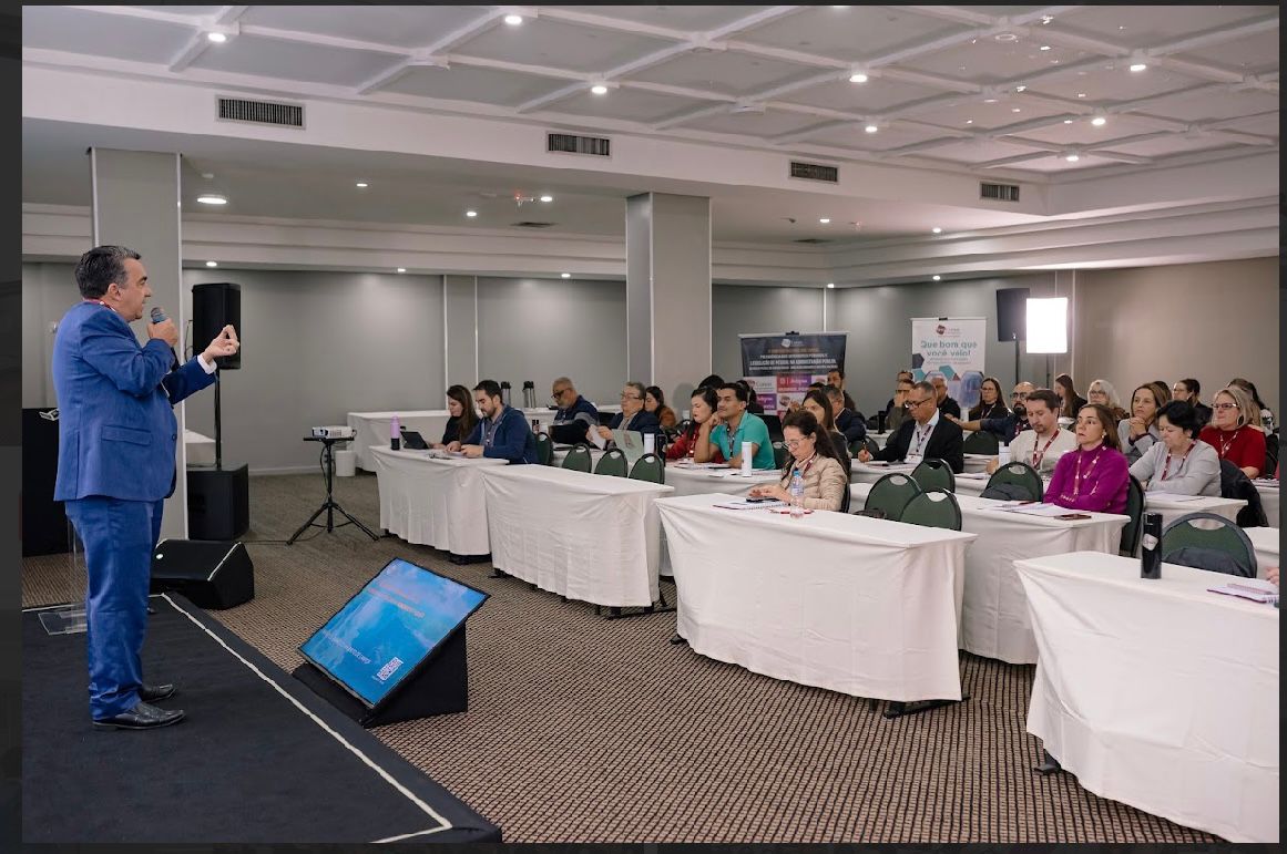 A imagem retrata o ambiente de um evento realizado em um amplo salão. Ao centro, um palestrante, vestido com terno azul, está em um pequeno palco elevado, conduzindo sua apresentação. À sua frente, uma plateia atenta ocupa várias filas de mesas retangulares dispostas em formato organizado, que preenchem a maior parte do espaço disponível. 