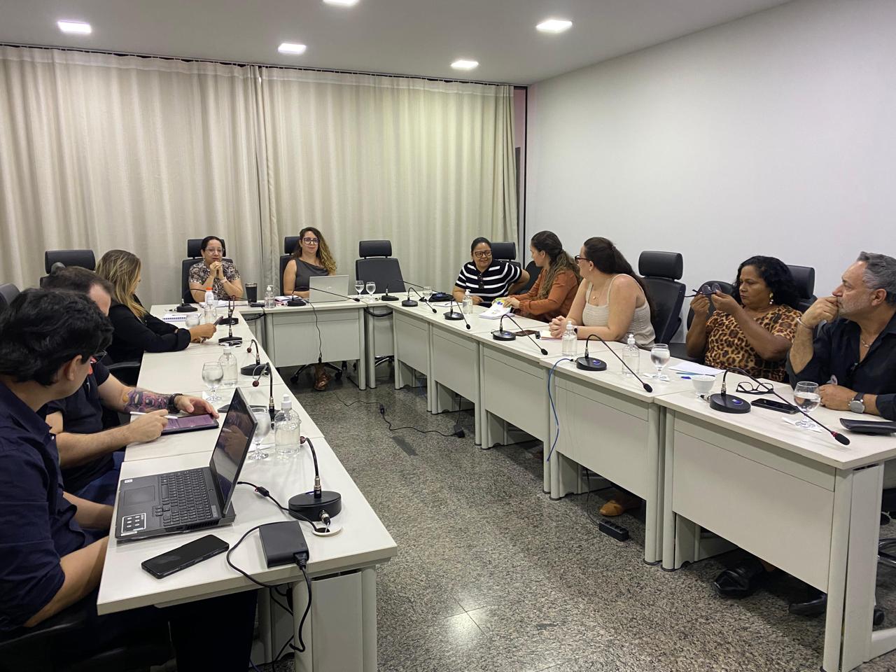 Homens e mulheres sentados em mesa branca em formato U, em sala de paredes brancas e persiana bege