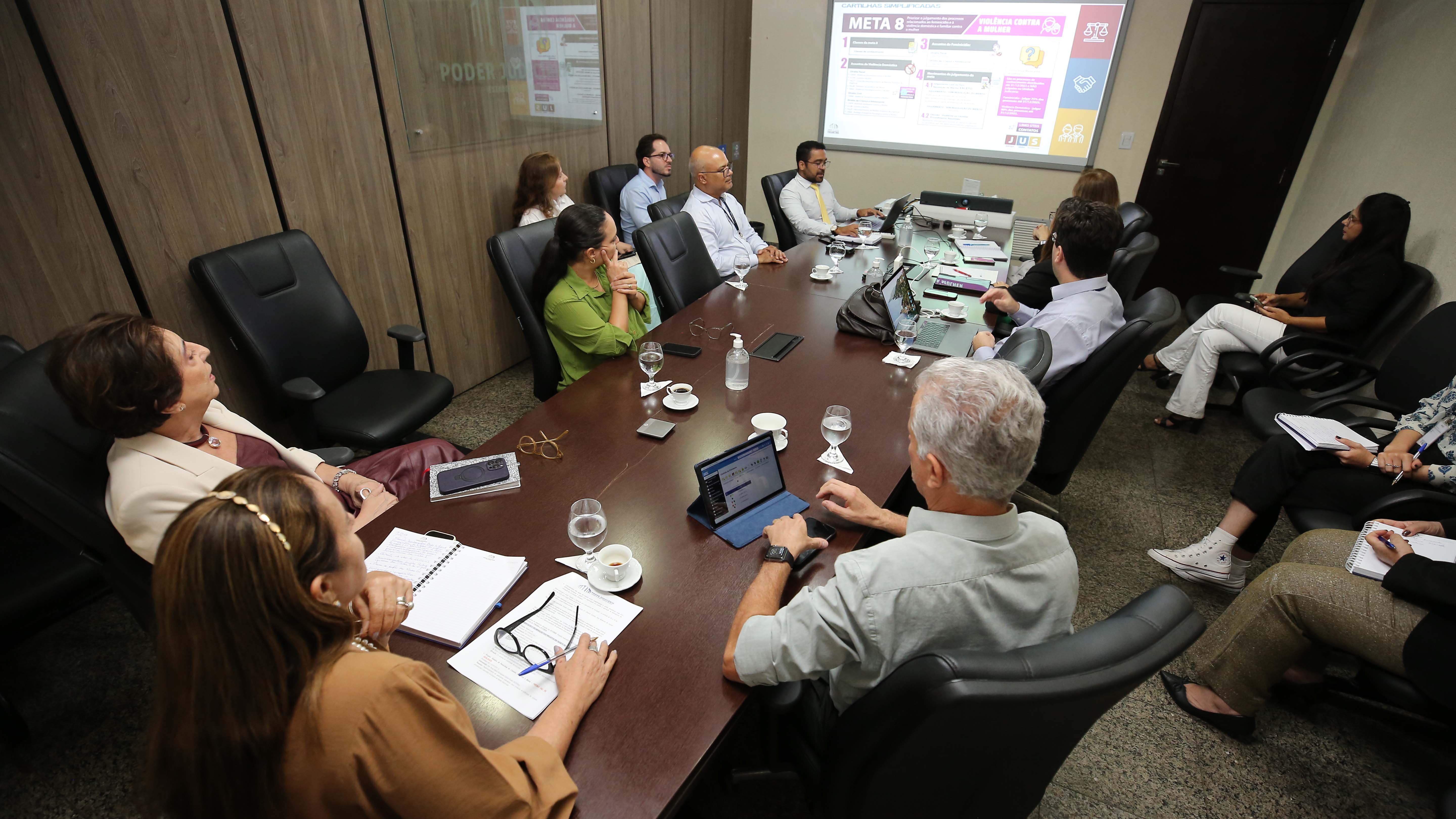 A imagem mostra um grupo de adultos reunidos em uma sala de reuniões, sentados ao redor de uma grande mesa retangular. Há diversidade de idade, aparência e trajes, que variam entre estilos profissionais e casuais. Sobre a mesa, há tablets e laptops.
