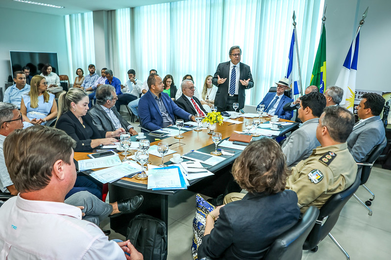 Reunião institucional realizada em uma sala com paredes claras e janelas cobertas por persianas verticais translúcidas. Ao redor de uma mesa oval estão cerca de 18 pessoas, entre elas homens e mulheres, sentados e em trajes formais. Um homem em pé, ao centro, fala aos presentes; atrás dele estão três bandeiras: do Brasil, do Tocantins e do Tribunal de Contas. Sobre a mesa há pastas, papéis, taças de água, flores amarelas e microfones. Ao fundo, outras pessoas acompanham a reunião sentadas. A atmosfera é formal e de articulação interinstitucional.