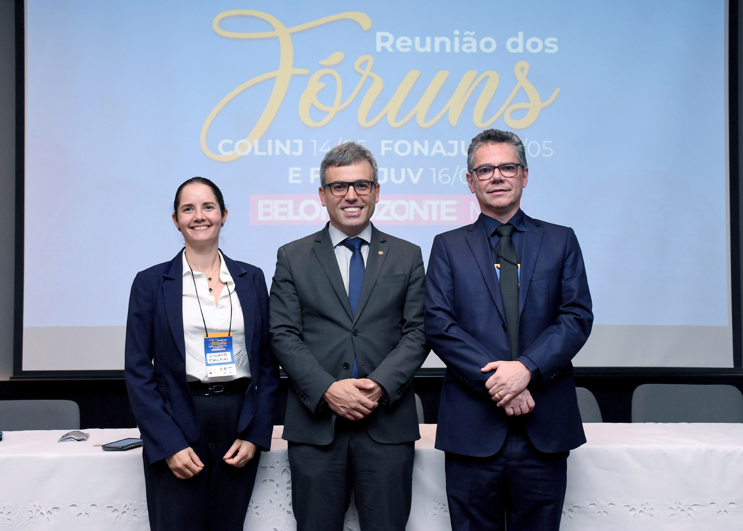 Três juízes, sendo dois homens e uma mulher, que está à esquerda, posam para foto durante evento, vestindo trajes formais.