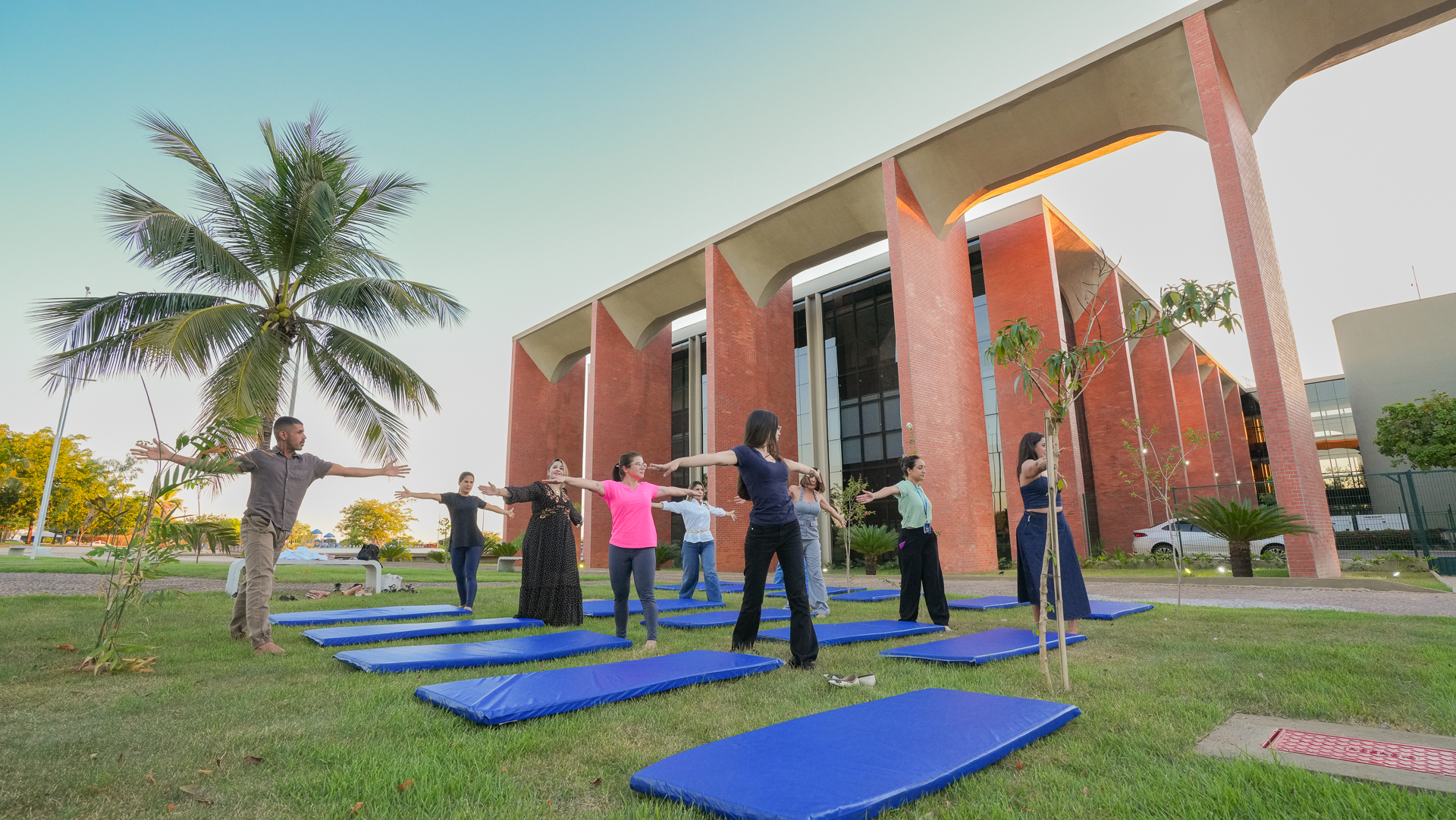 Várias pessoas praticando yoga no gramado em frente ao prédio do Tribunal de Justiça, na imagem aparecem tapetes azuis e uma palmeira à esquerda