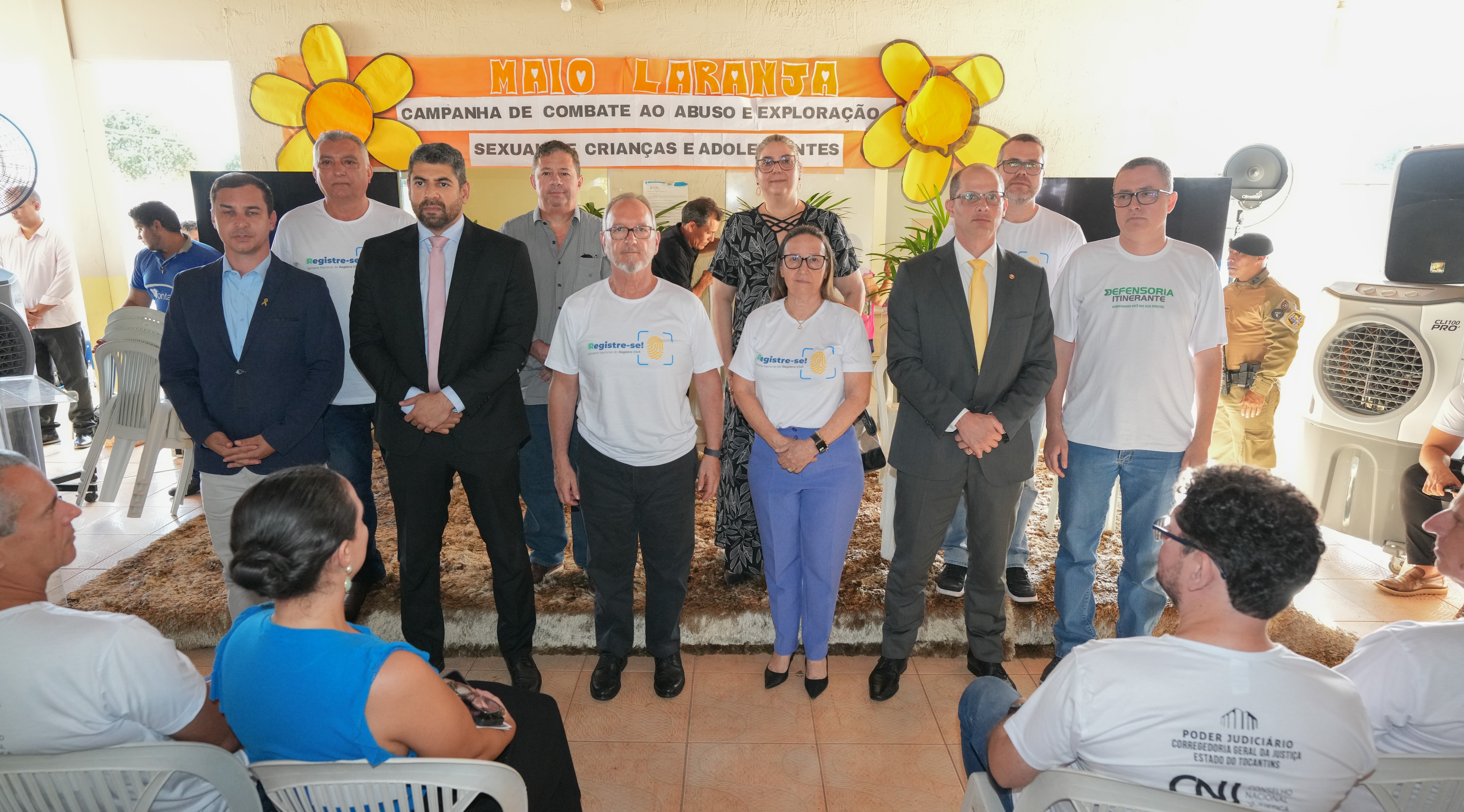 Dez autoridades e representantes institucionais posam em duas fileiras sobre um tapete marrom, voltados para a câmera, enquanto participantes sentados ocupam as cadeiras em primeiro plano. Ao fundo, um painel laranja traz o título “Maio Laranja – Campanha de Combate ao Abuso e Exploração Sexual de Crianças e Adolescentes”, ladeado por flores amarelas de papel. Na fileira da frente, dois homens de terno escuro e gravata dividem espaço com três pessoas vestindo camisetas brancas do projeto “Registre‑se” e uma mulher de blusa branca e calça azul; atrás deles, outros cinco participantes completam a composição. Equipamentos de som, ventiladores e um policial militar à direita reforçam o clima institucional da cerimônia.