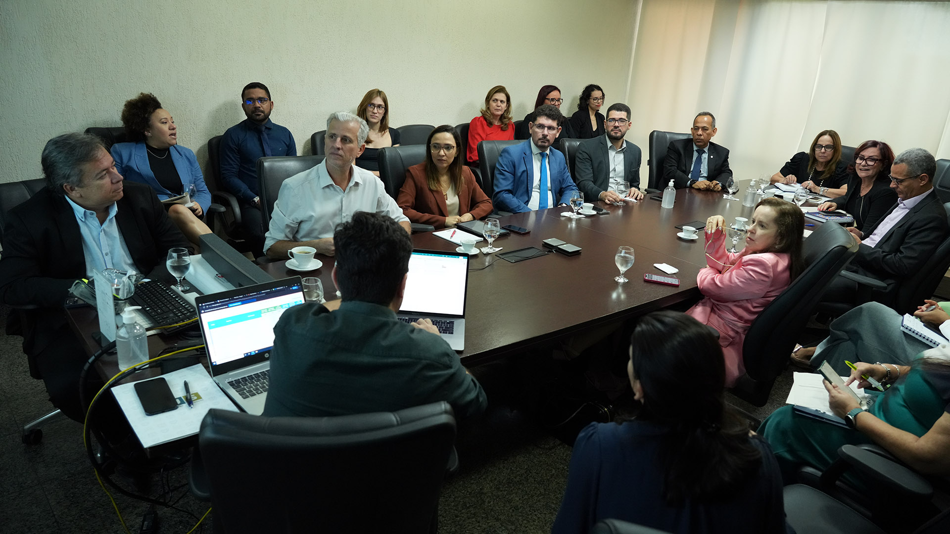 Várias pessoas reunidas em uma sala, todas sentadas. Algumas em volta de uma grande mesa de reunião.  