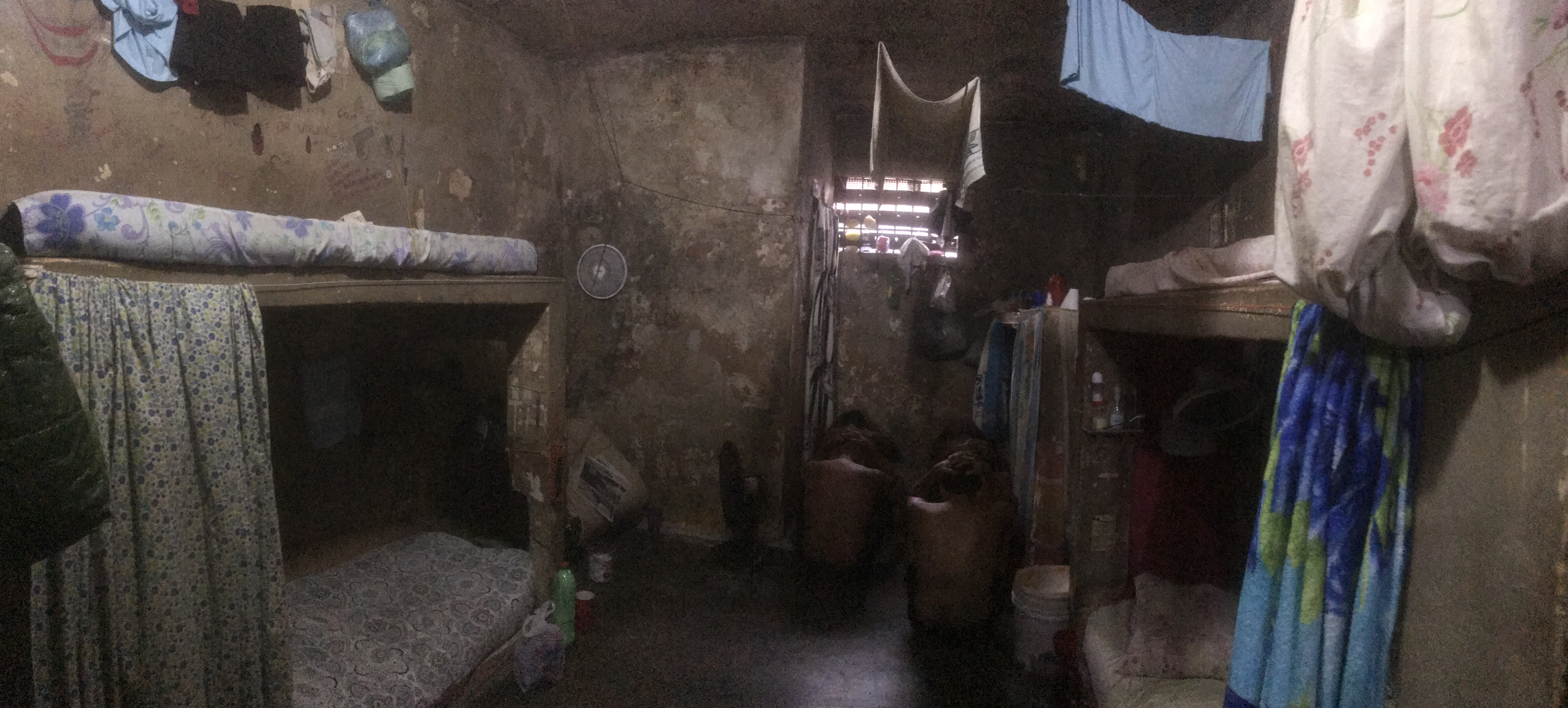 Imagem de uma cela em condições precárias com internos de costas, sentados no chão.