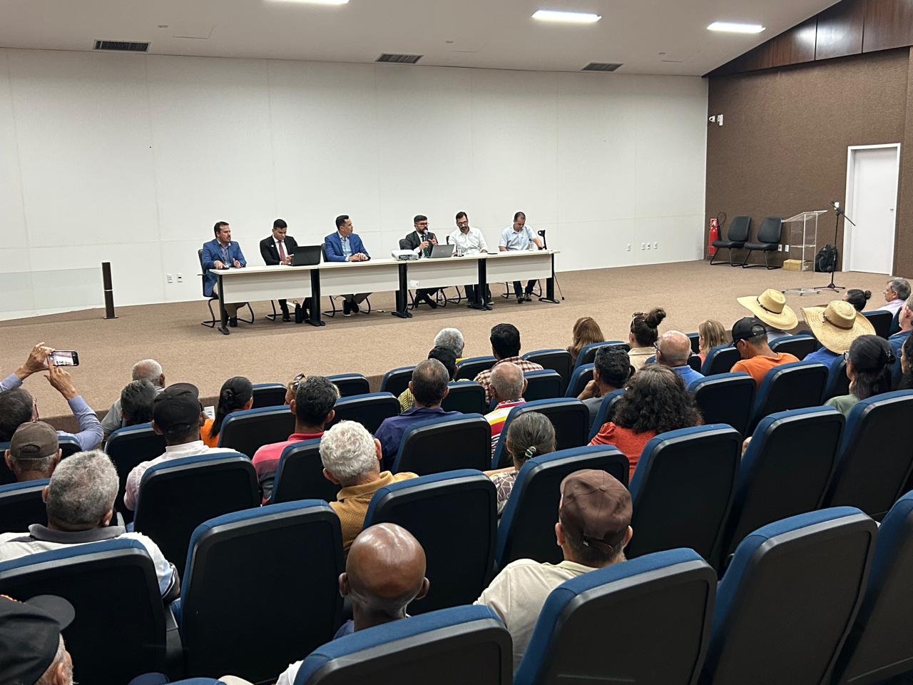 Imagem mostra um auditório com várias pessoas sentadas em cadeiras azuis; ao fundo, no palco, uma mesa com seis homens sentados de frente para o público e ao centro o juiz fala ao microfone.