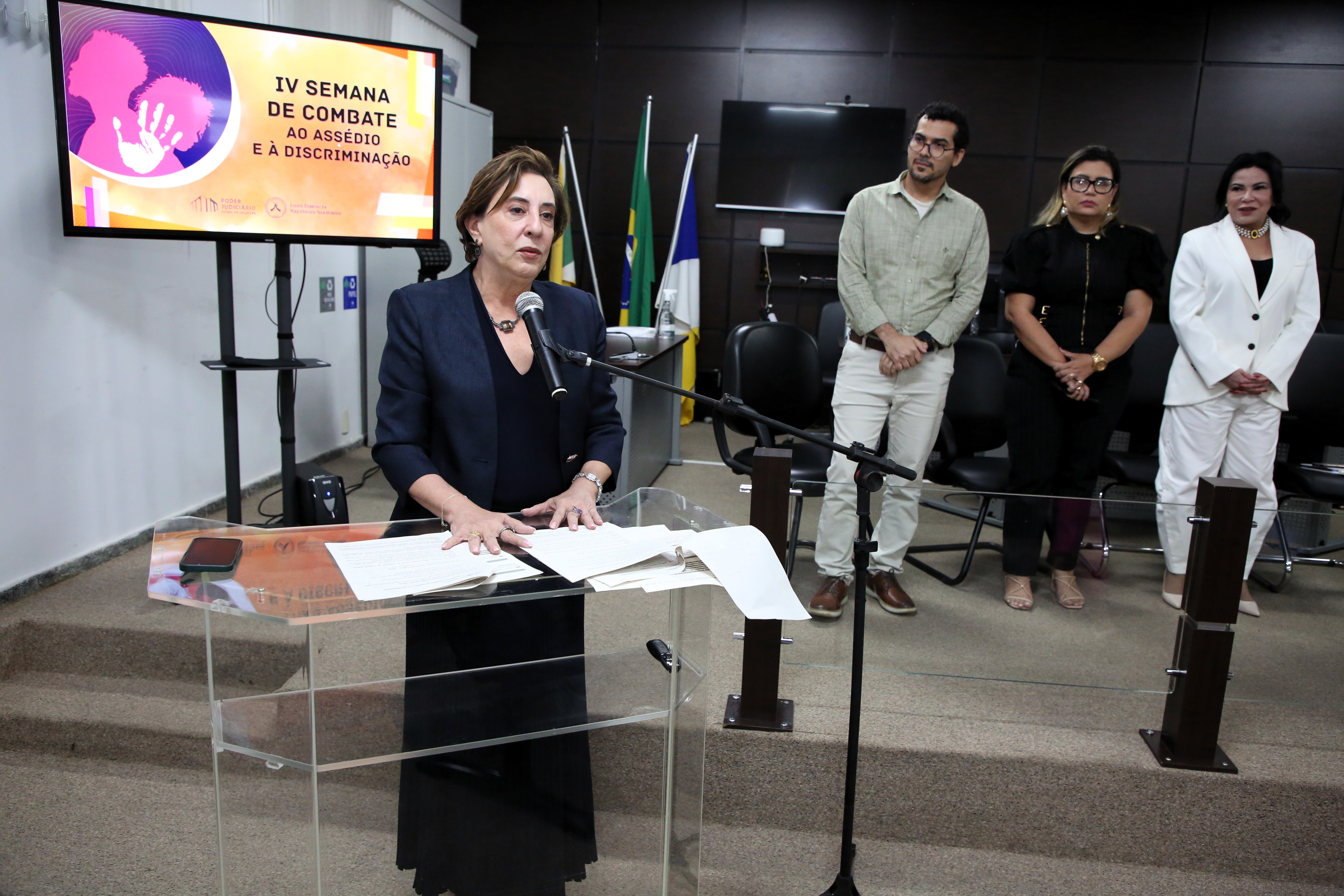 imagem mostra a desembargadora Angela Haonat no púlpito, falando ao microfone, com outras pessoas presentes.
