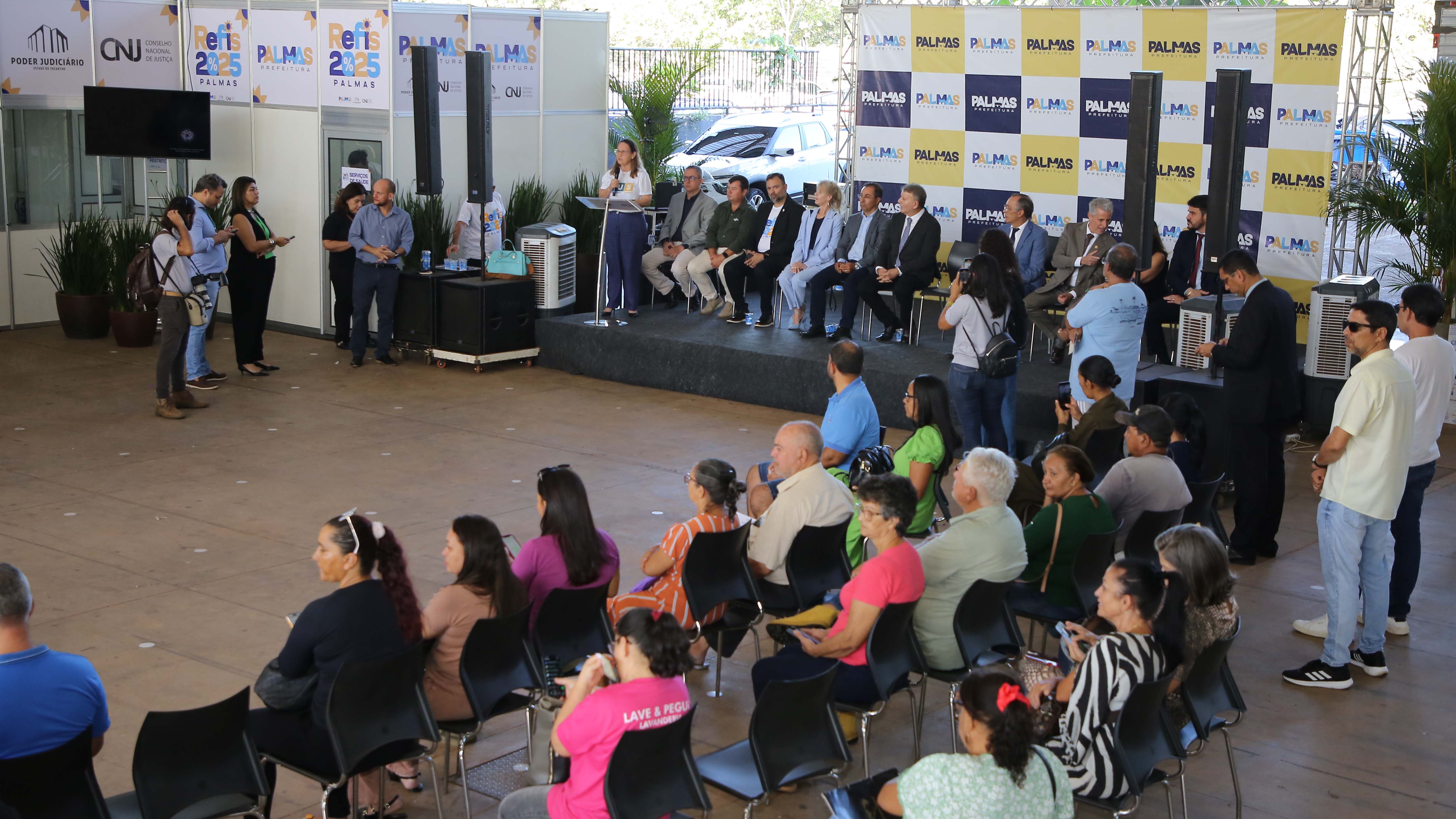 Homens e mulheres em sentados assistindo mulher branca de cabelos lisos e soltos, falando ao microfone em púlpito em palco com autoridades sentadas durante abertura de evento de renegociação fiscal