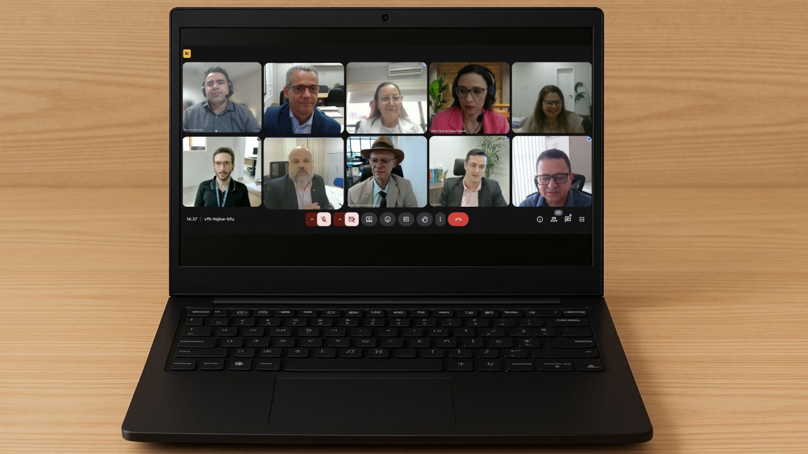 Notebook sobre uma mesa de madeira, exibindo uma videoconferência na tela. A reunião virtual reúne dez pessoas, distribuídas em dois blocos de cinco participantes cada. Todos aparecem em seus respectivos quadrados, com expressões atentas, em ambientes internos, alguns com fundo de escritório, outros com parede branca ou decoração ao fundo. A interface da videoconferência mostra os ícones tradicionais de microfone, câmera, compartilhar tela e encerrar chamada na parte inferior. A tela está centralizada, e o ambiente ao redor é neutro, focando na tela do computador.
