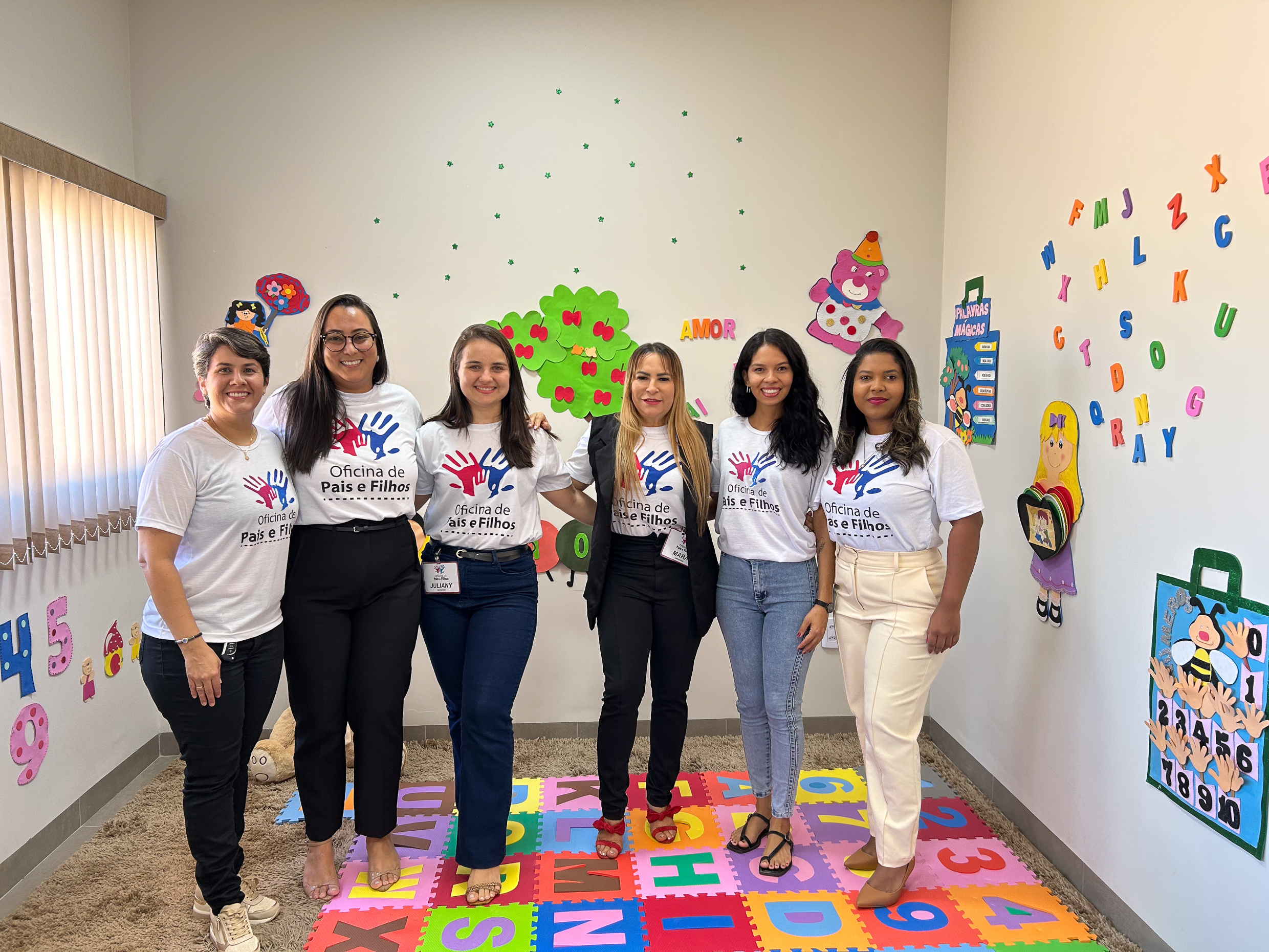 Seis mulheres estão em pé, lado a lado, sorrindo para a foto em uma sala decorada com elementos infantis. Todas vestem camisetas brancas com a estampa “Oficina de Pais e Filhos”, que traz o desenho de duas mãos coloridas (uma azul e uma vermelha). Algumas usam calças jeans e outras calças sociais, e duas delas também vestem blazer. O chão é coberto por tapetes coloridos de EVA com letras e números, e as paredes estão enfeitadas com figuras de animais, árvore com corações e desenhos infantis. A palavra “AMOR” está em destaque na parede ao fundo. A iluminação natural entra por uma janela com persiana à esquerda.