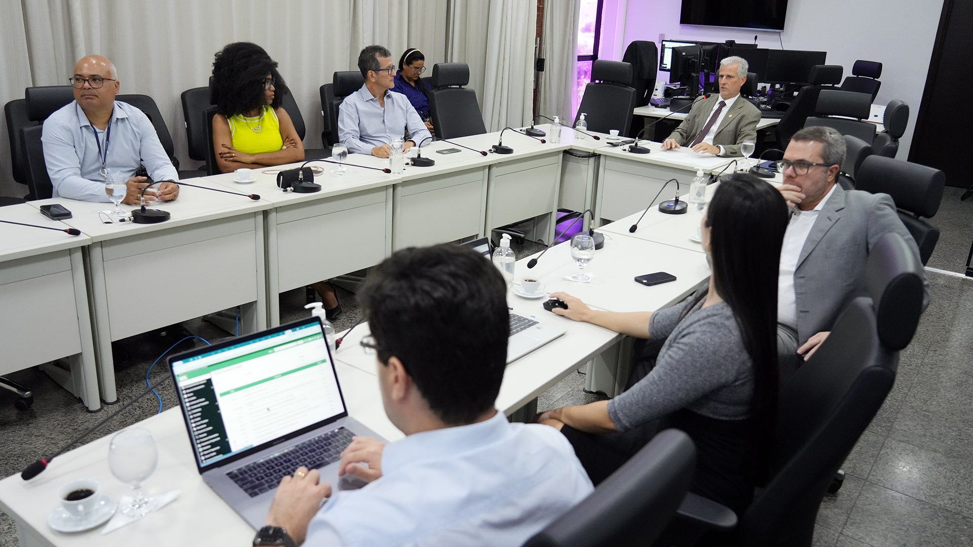 Reunião em sala ampla e bem iluminada do TJTO. Nove pessoas estão sentadas em torno de uma mesa branca em formato de “U”. Elas observam, escutam ou usam notebooks. À frente, microfones, copos com água e xícaras de café. Ao fundo, cortinas claras e telas de monitoramento ligadas.