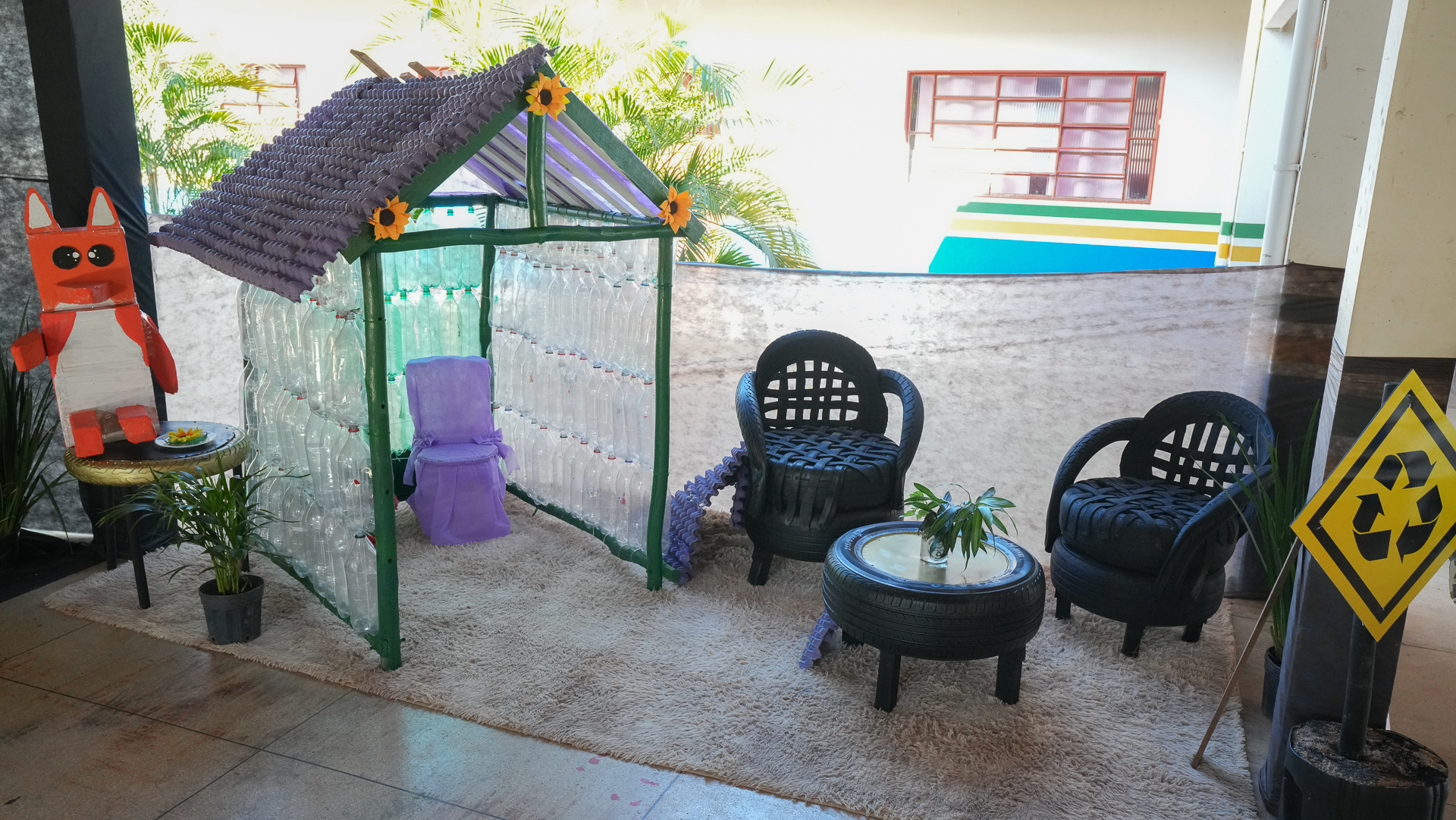 A foto mostra um ambiente decorado com materiais recicláveis, como garrafas PET, pneus e caixas de ovos. O destaque é uma casinha feita com garrafas e telhado roxo, além de móveis como poltronas e mesa produzidos com pneus. O espaço é complementado com plantas, um boneco colorido e um tapete, transmitindo uma proposta criativa de sustentabilidade e educação ambiental.