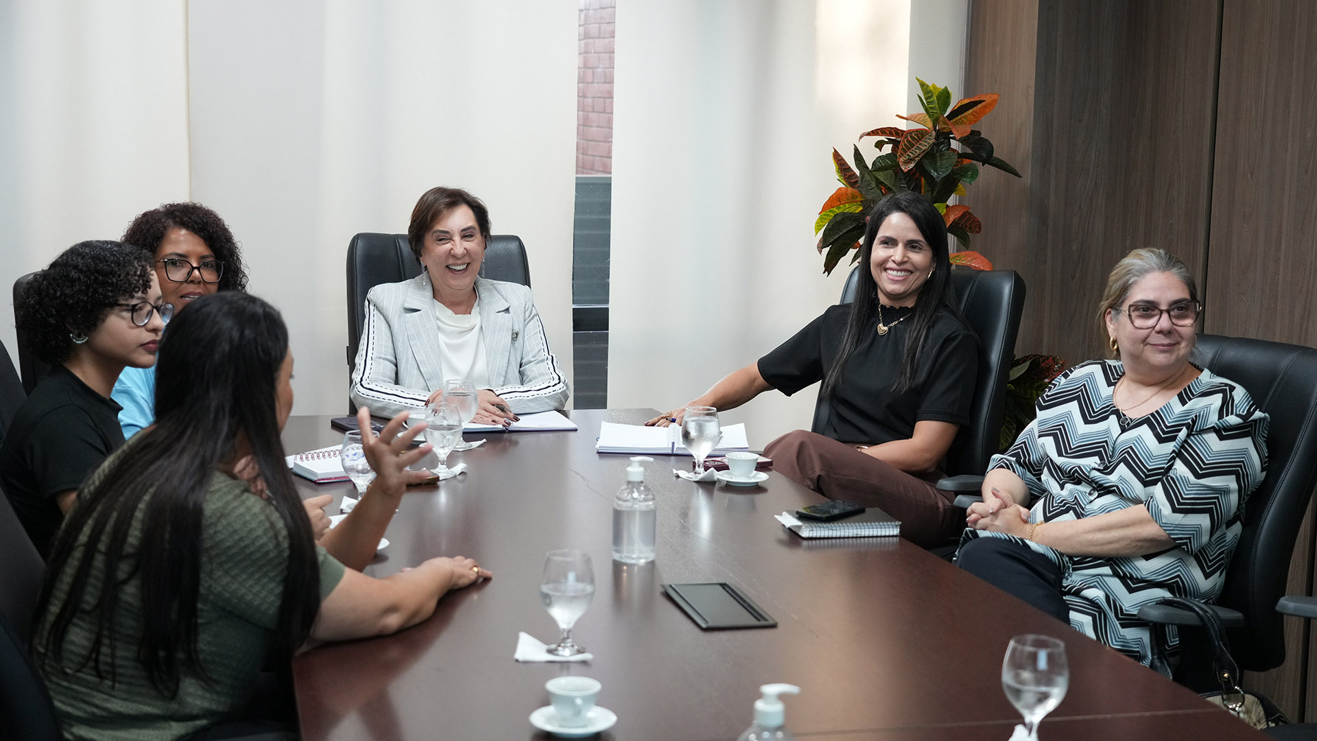 Homens e mulheres sentados em mesa branca em formato U, em sala de paredes brancas e persiana bege