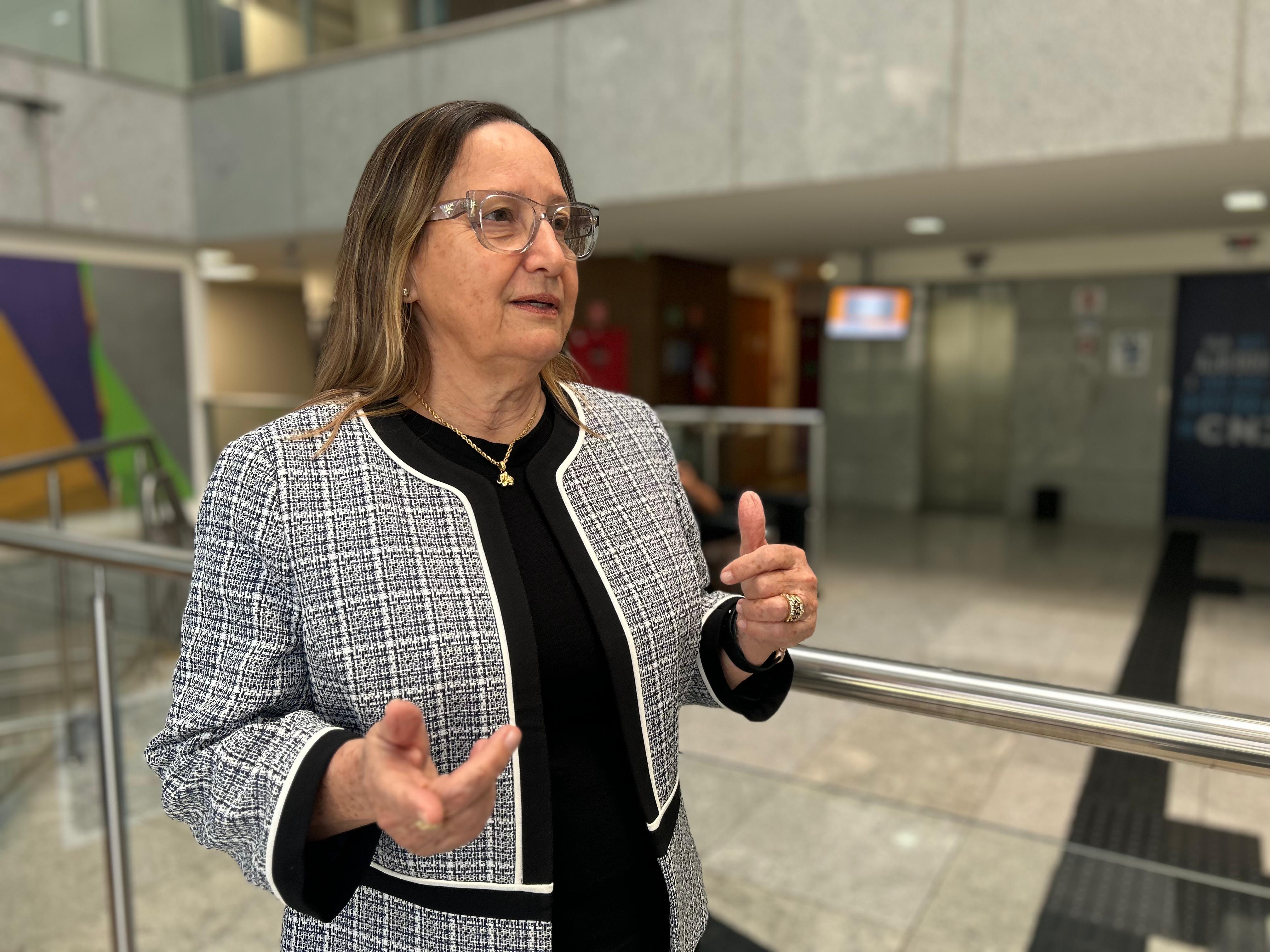 A presidente do Tribunal de Justiça do Tocantins, desembargadora Maysa Vendramini Rosal, aparece em primeiro plano, gesticulando com as mãos enquanto fala. Ela veste blazer com estampa xadrez em preto e branco, blusa preta por baixo e usa óculos transparentes. Ao fundo, o interior moderno de um prédio institucional, com paredes claras, corrimão de inox e ambiente corporativo.