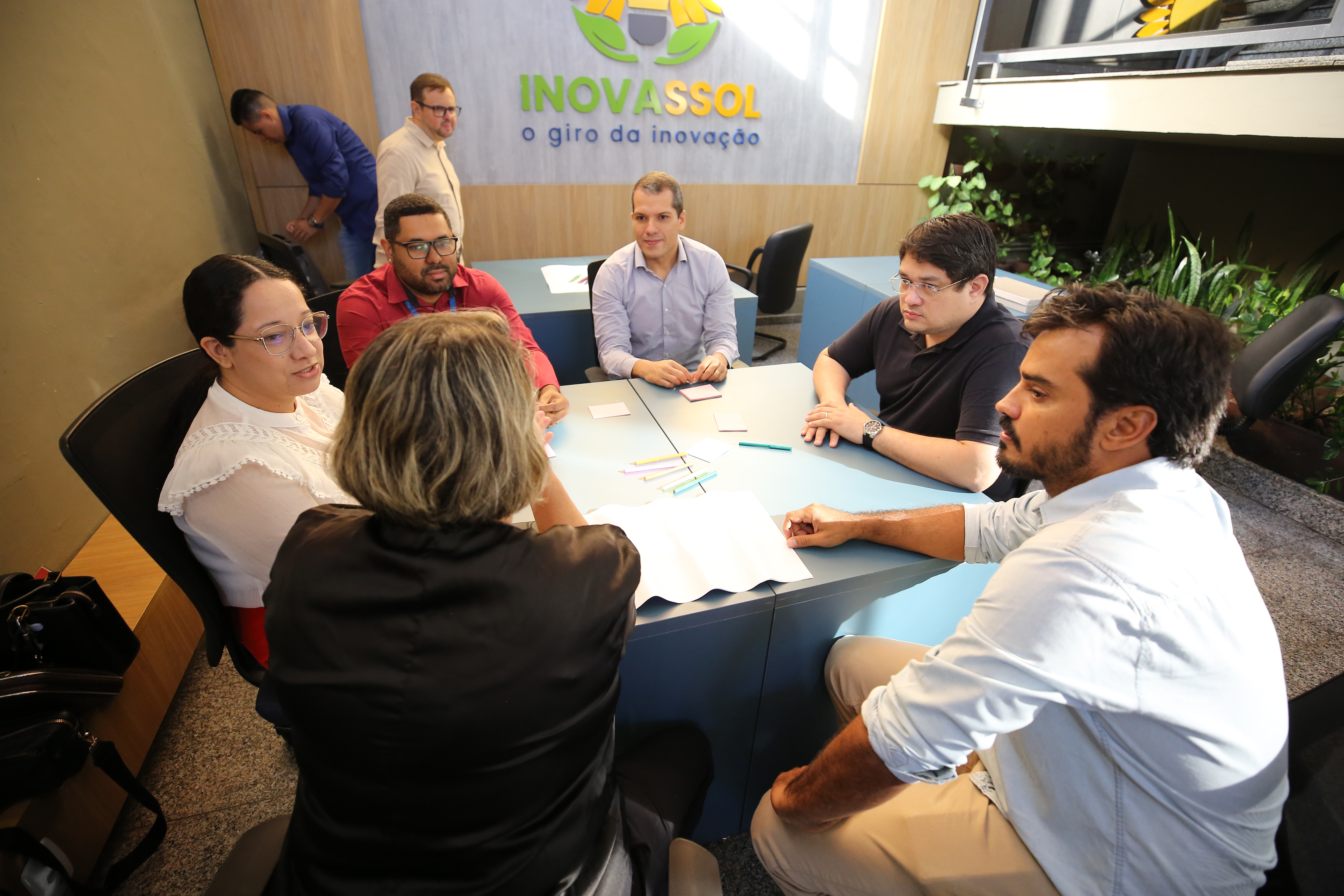 Na imagem aparece seis pessoas estão reunidas em torno de uma mesa azul clara, em ambiente interno, participando de uma roda de conversa ou dinâmica de grupo. Sobre a mesa há papéis em branco e canetas coloridas. Ao fundo, na parede, há um painel com a identidade visual do projeto “INOVASSOL – o giro da inovação”. Duas outras pessoas estão ao fundo, uma em pé próxima à parede e outra ao lado do painel. O local é iluminado, com decoração moderna e presença de plantas ornamentais.